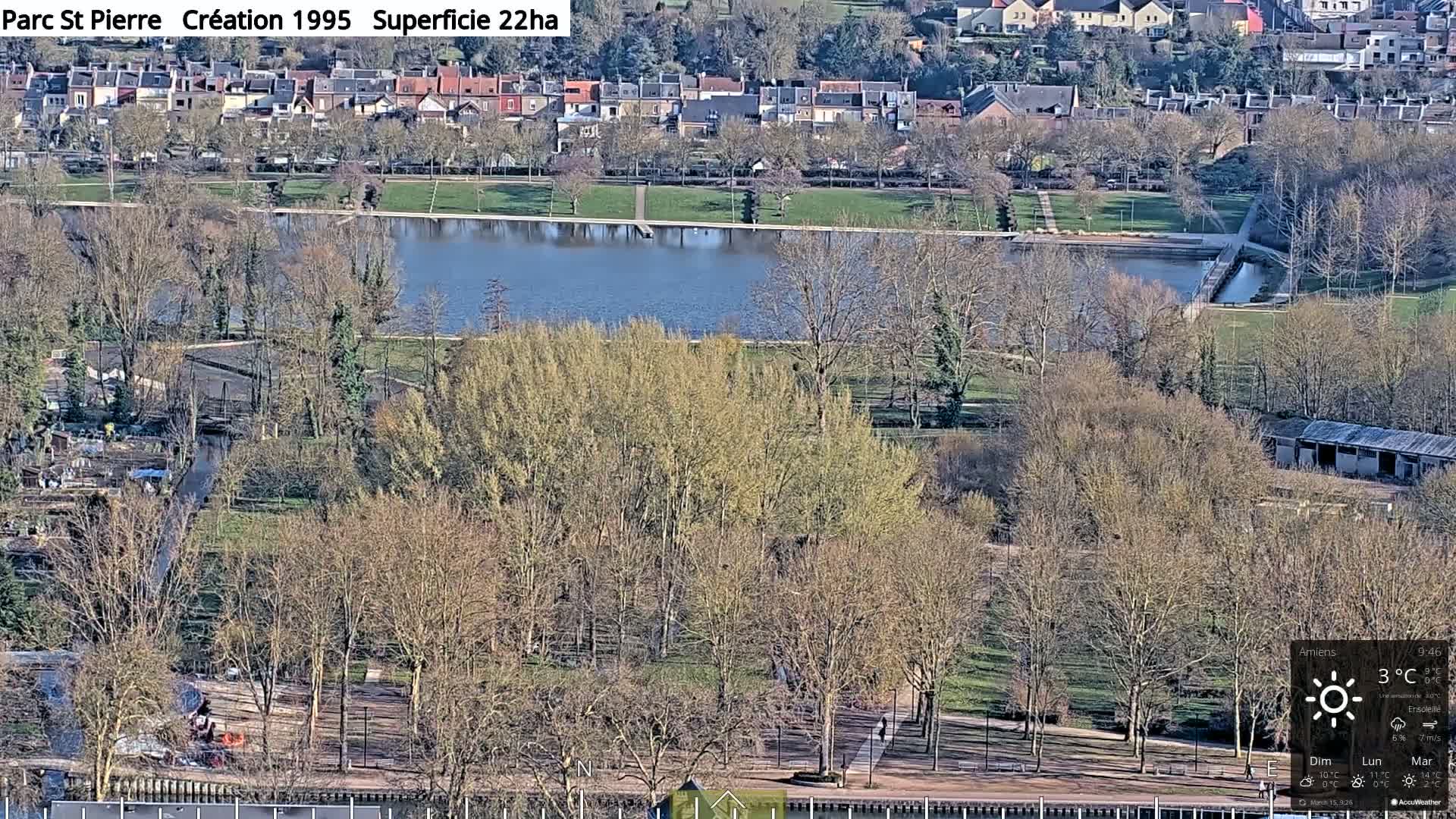 An aerial view of a park with a pond, bare trees, and residential buildings in the background on a sunny day.