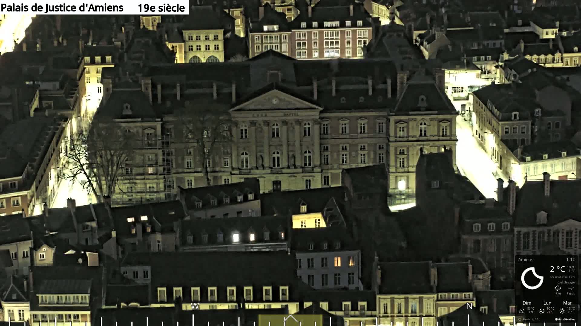 A nighttime aerial view of a city block showing a large, brightly lit building surrounded by smaller, darker buildings under a clear sky.