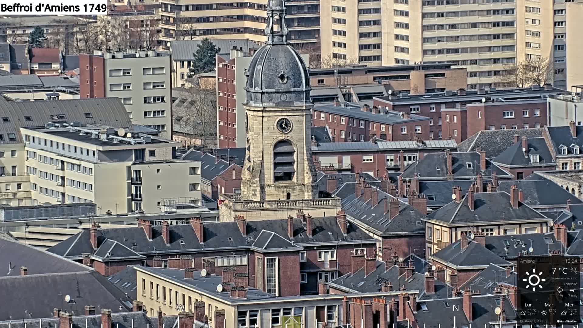 A tall clock tower stands in the center of a city under a sunny sky, surrounded by many closely packed buildings with dark, pitched roofs.