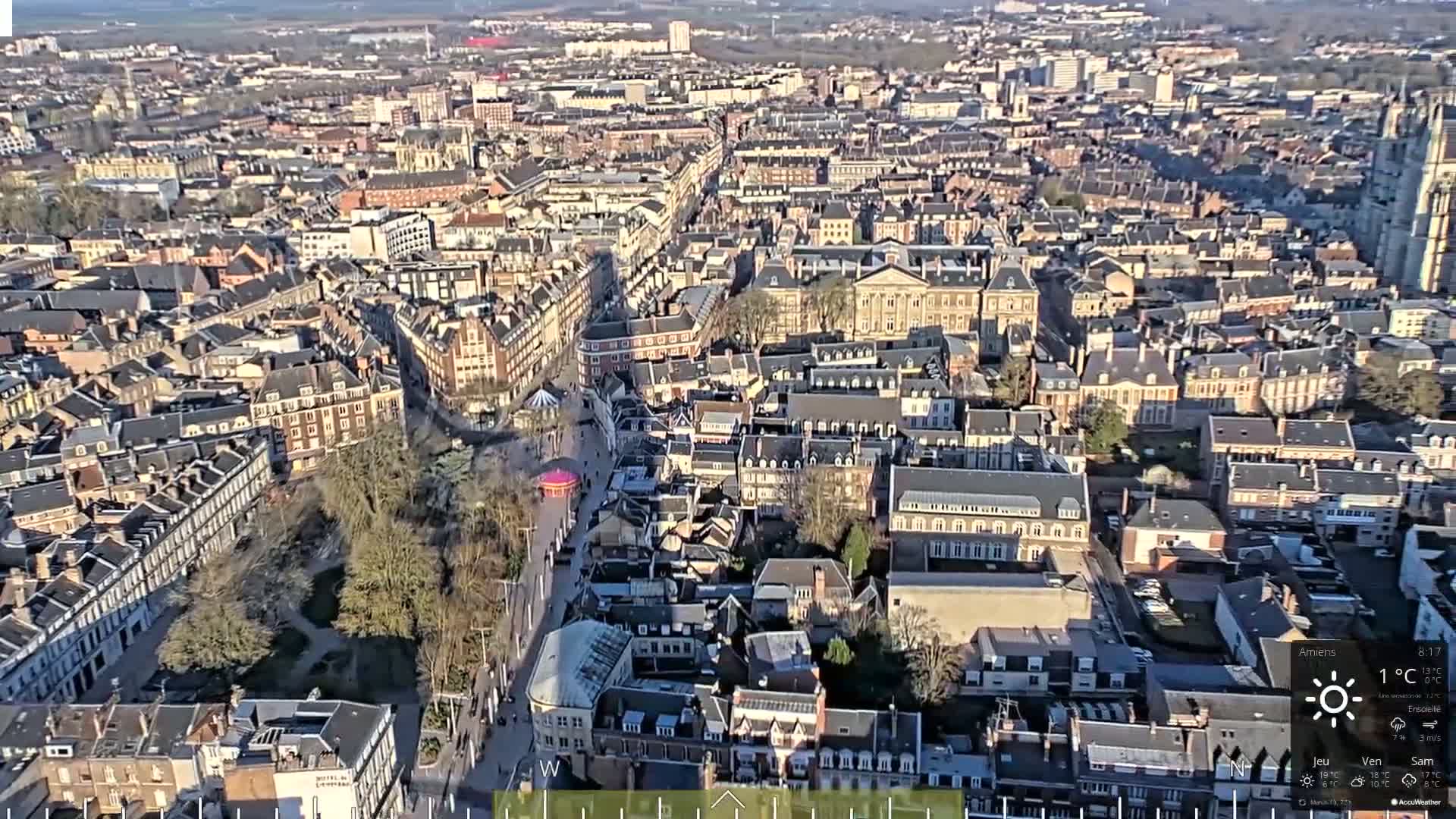 A high-angle, sunny view of a densely packed European city with primarily dark-roofed buildings, a central street, and a small park.