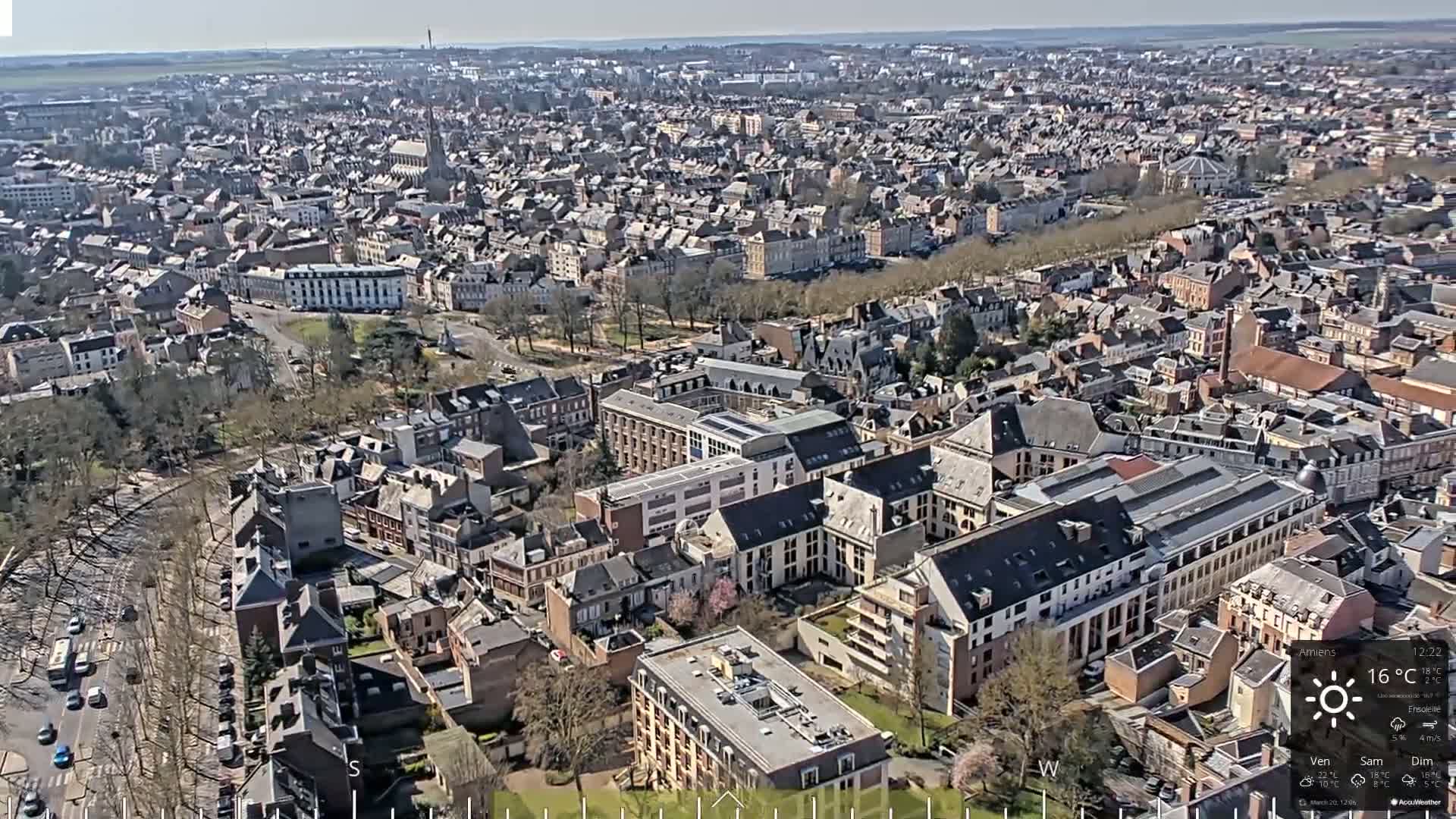 A high-angle, sunny view of a densely populated city with numerous buildings of varying sizes and styles, surrounded by trees and green spaces.