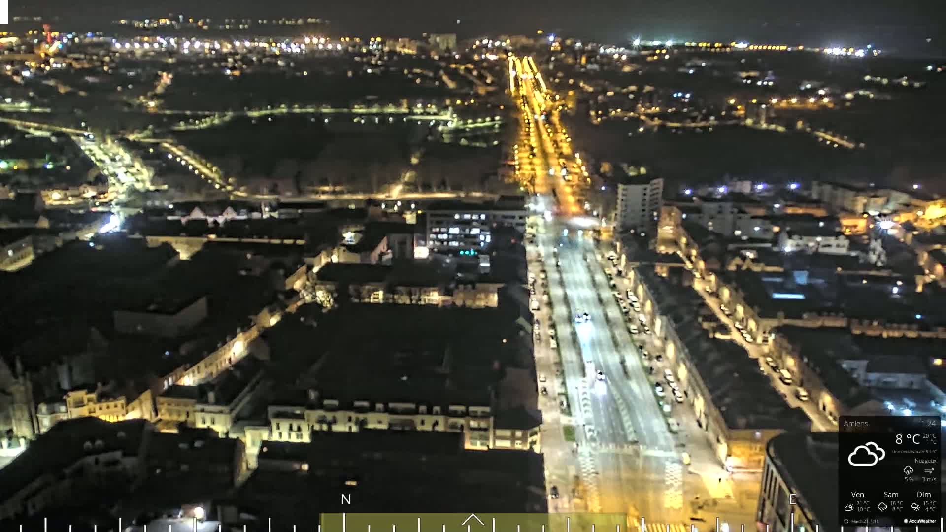 A nighttime, high-angle view of a city with a long, brightly lit street running through it, under cloudy skies.