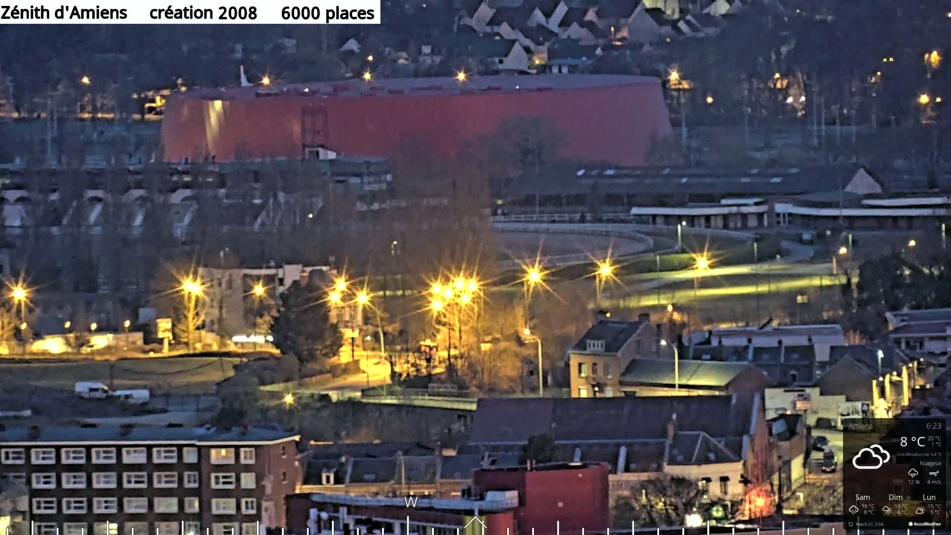 A nighttime aerial view of a city with a large red building, illuminated streets, and a lit-up oval track, under cloudy skies with a temperature of 8°C.