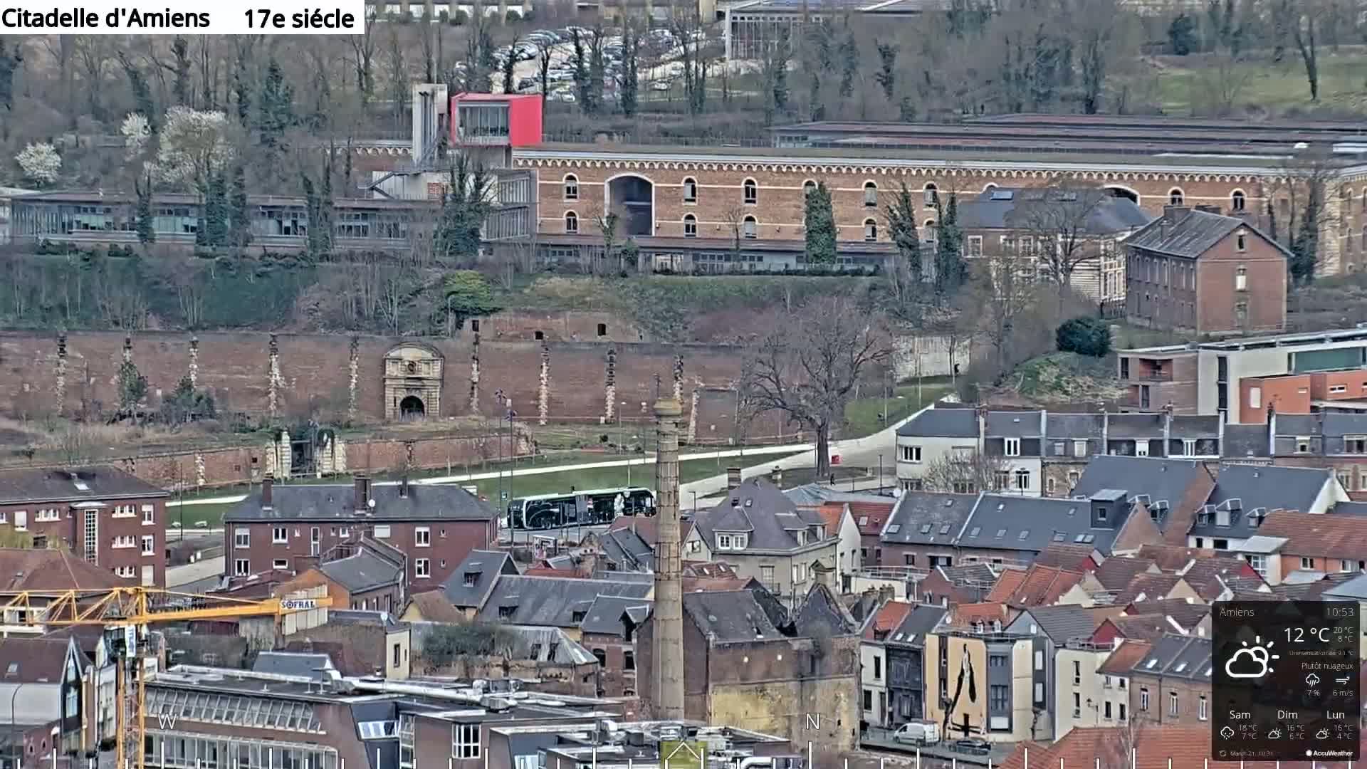 A high-angle, long shot of a European city on a partly cloudy day, featuring a large brick structure and a tall industrial chimney, with a mix of residential buildings and a construction crane visible below.