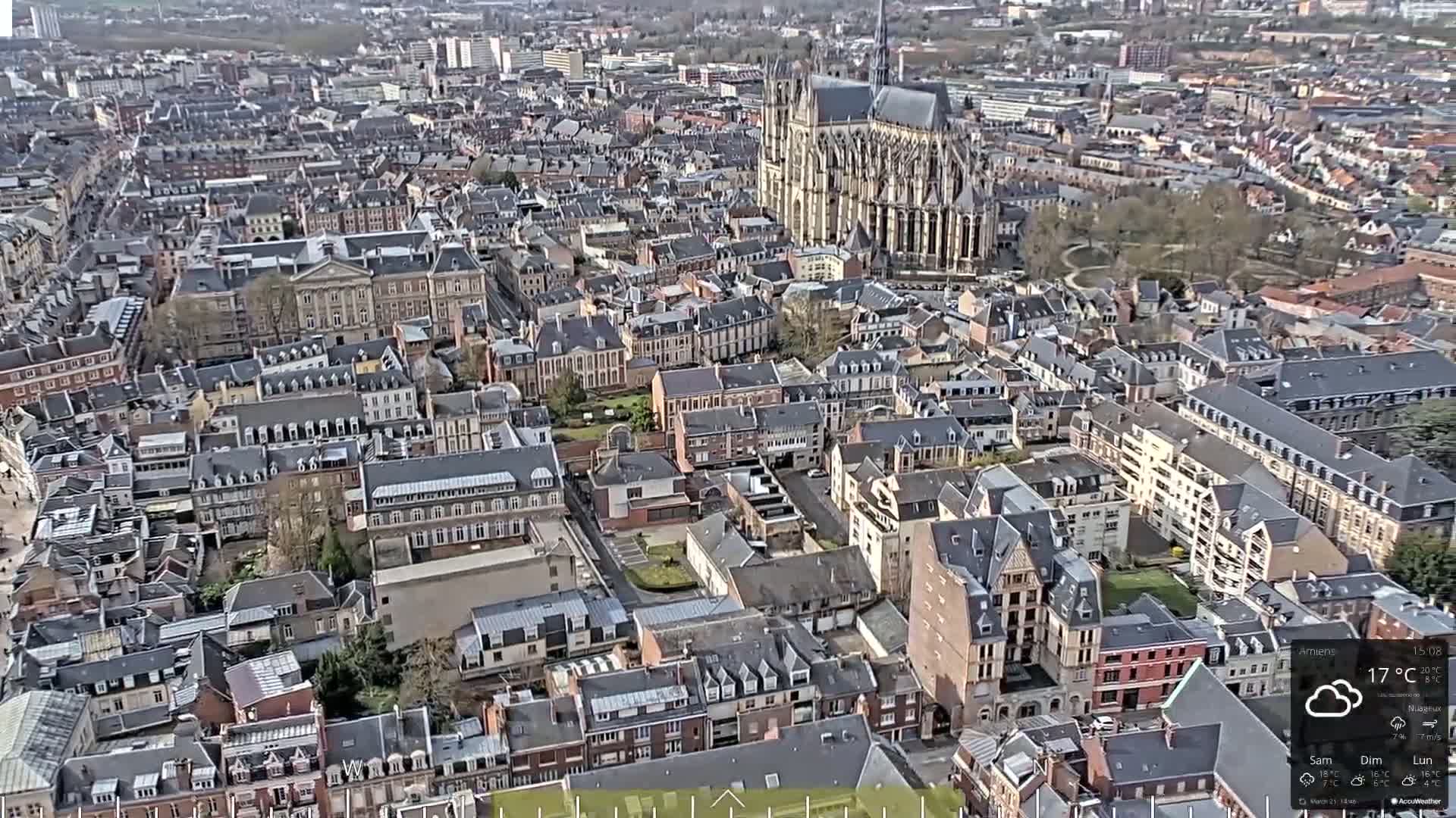 An aerial view of a European city on a partly cloudy day shows a large cathedral and densely packed buildings.