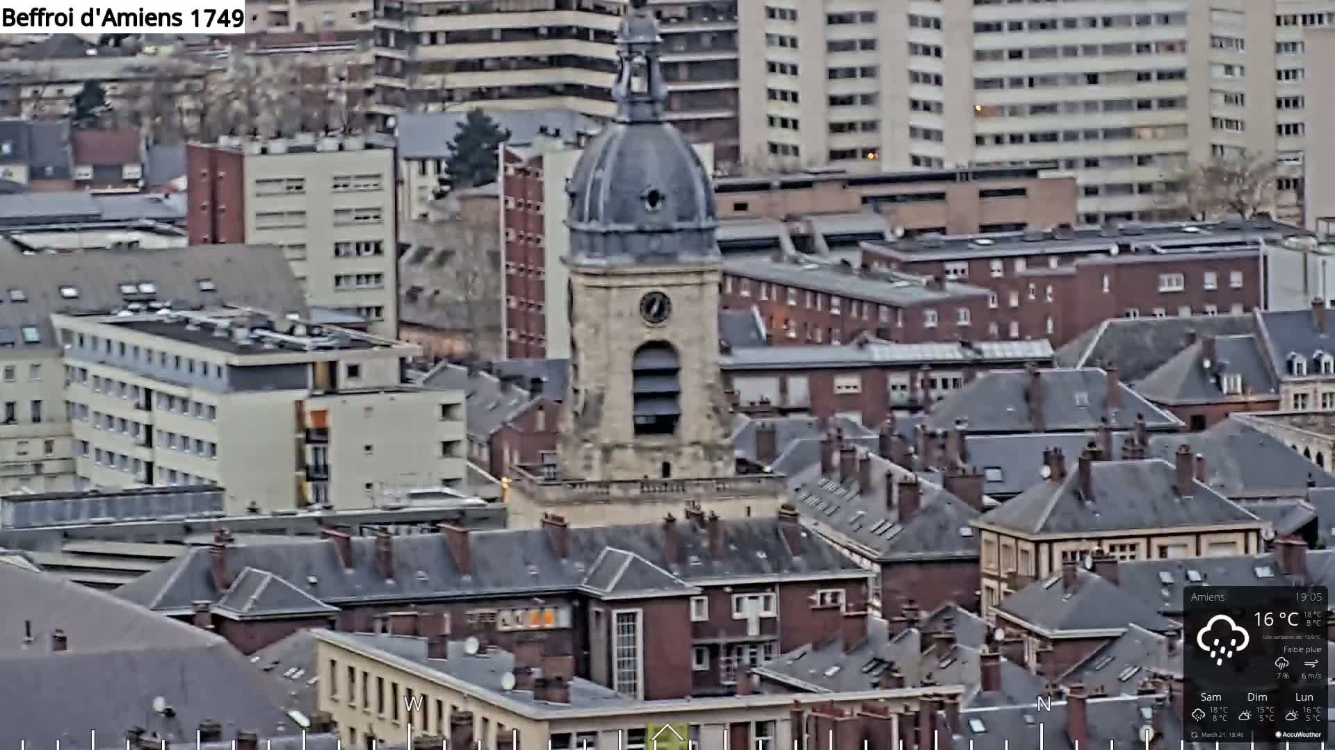 A high-angle, long shot of a European city on an overcast day with light rain shows a large clock tower surrounded by many buildings with dark grey slate roofs.