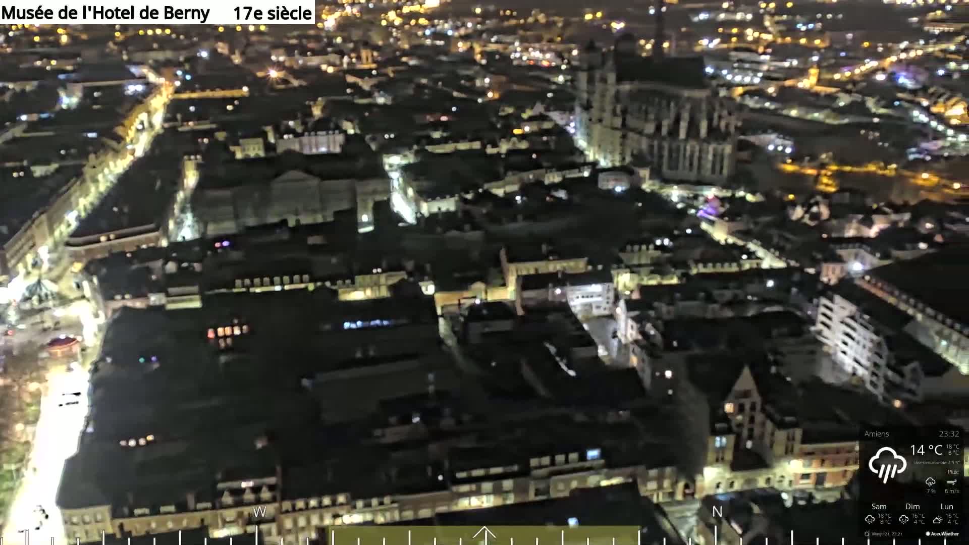 A nighttime aerial view of a city with many illuminated buildings, streets, and a large cathedral-like structure under cloudy conditions with rain.