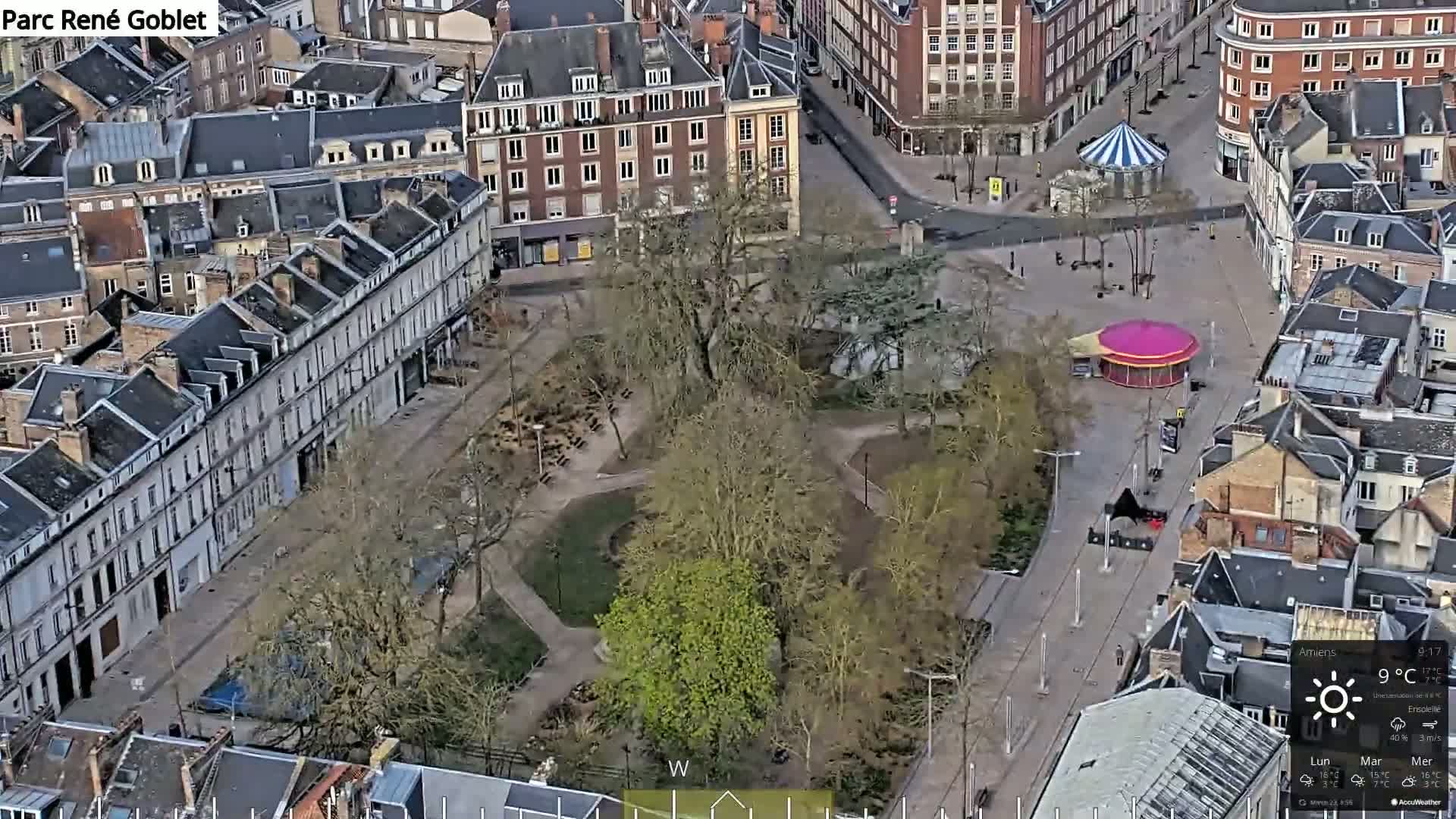 An aerial view shows a city square with a park in the center, surrounded by buildings, on a sunny day with a temperature of 9°C.
