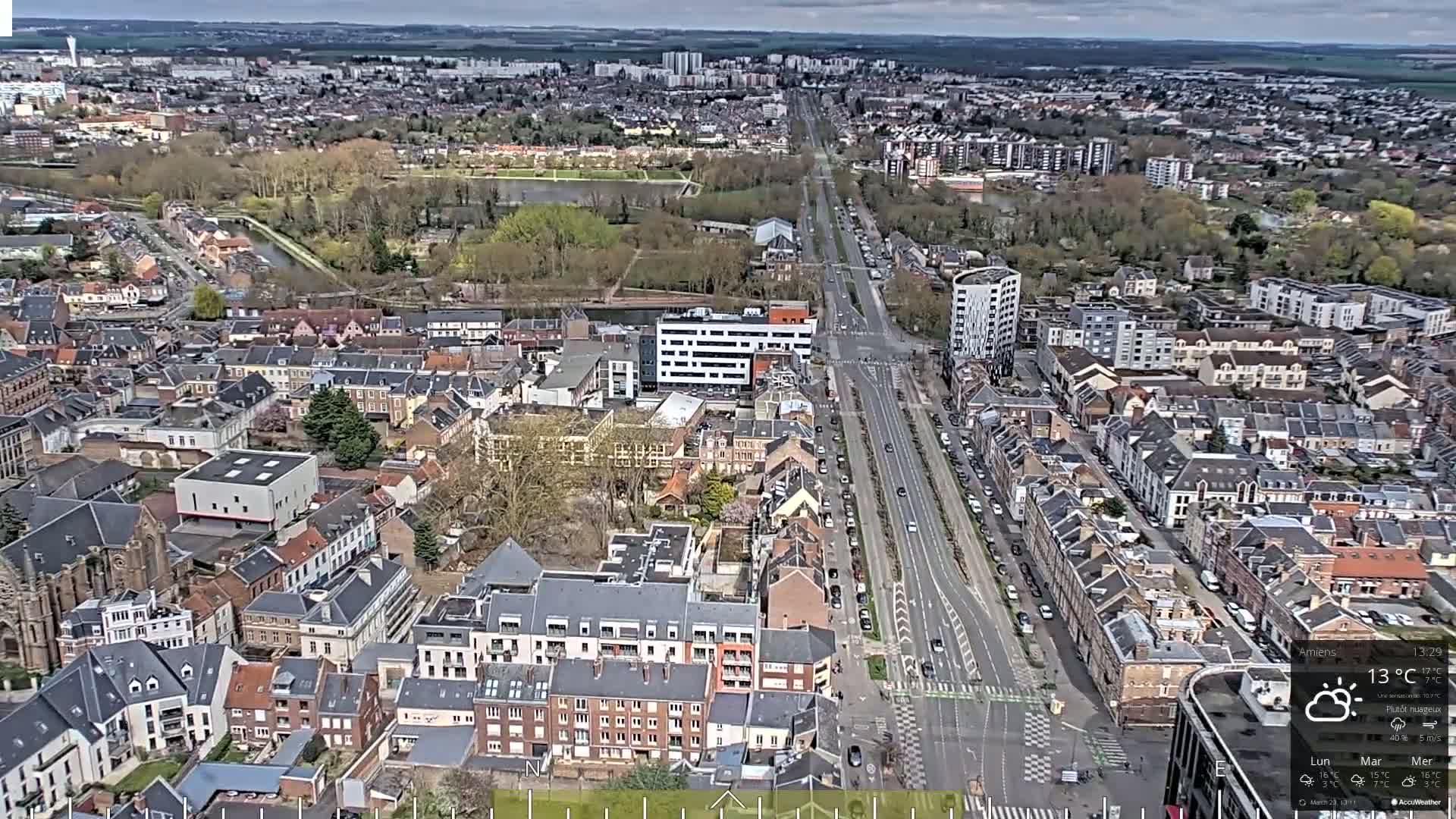 An aerial view of a city on a partly cloudy day shows a long road flanked by residential buildings, with a wide expanse of greenery and a body of water visible in the distance.