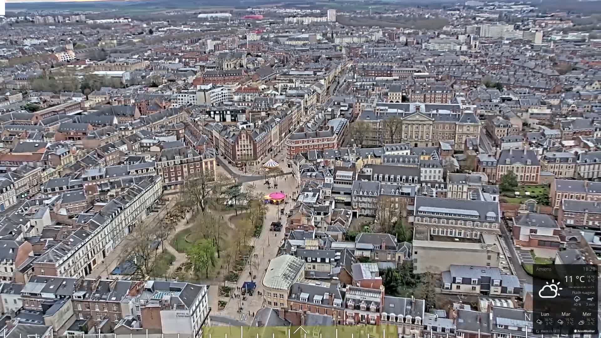 A high-angle, long shot of a city on a partly cloudy day shows numerous buildings and a park in the center of the city.