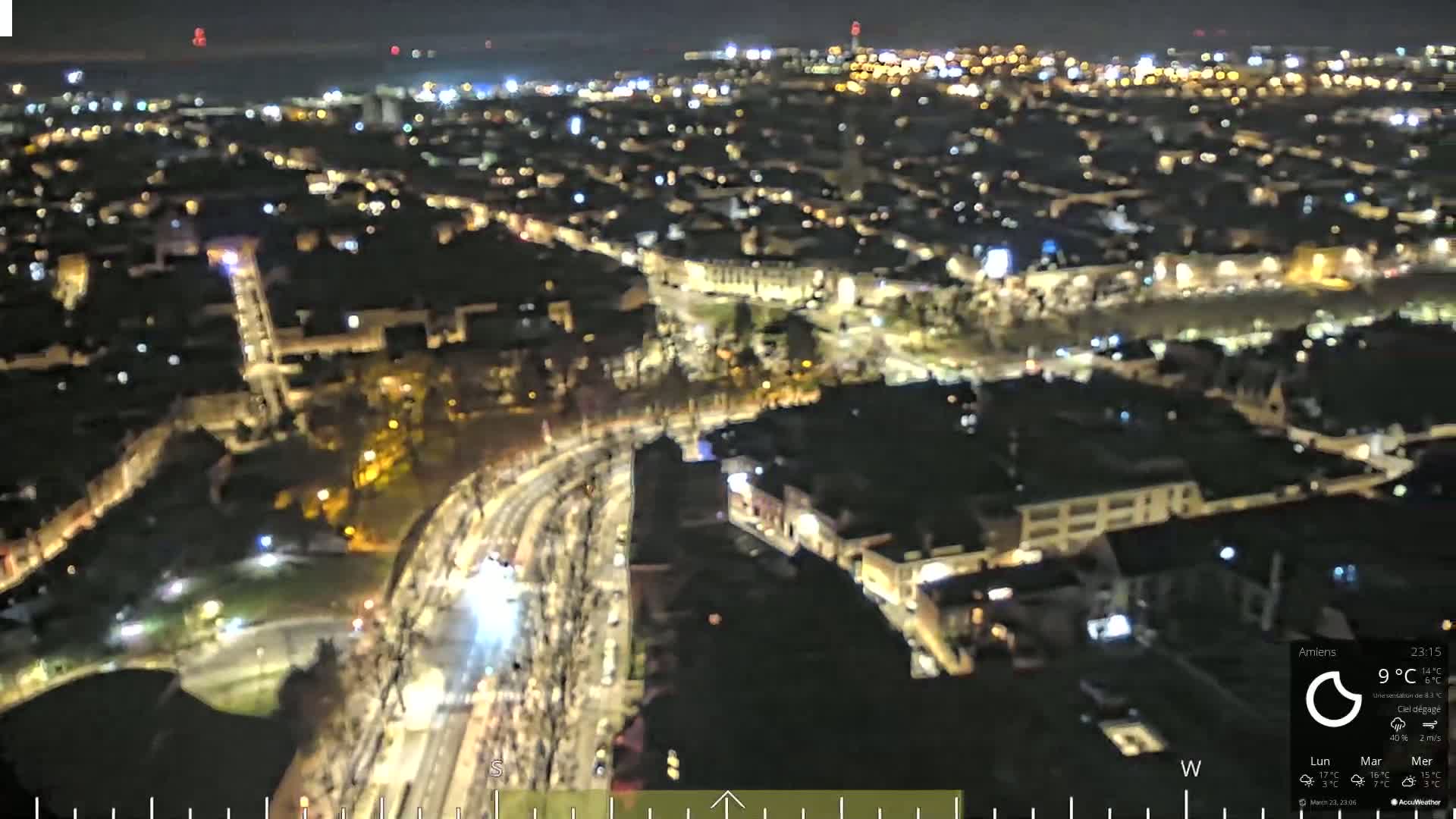 A nighttime, aerial view of a city with brightly lit streets and buildings, clear skies, and a large gathering of people along a road.