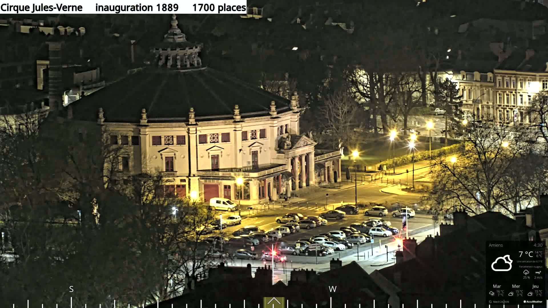 A nighttime aerial view of a round, illuminated building with a parking lot in front, surrounded by bare trees and streetlights under partly cloudy skies.