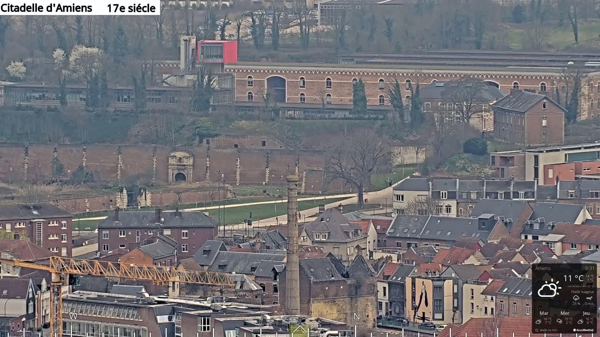 A high-angle, long shot of a city with mostly overcast skies shows a brick citadel wall and gateway, a tall industrial chimney, many buildings, and a construction crane.