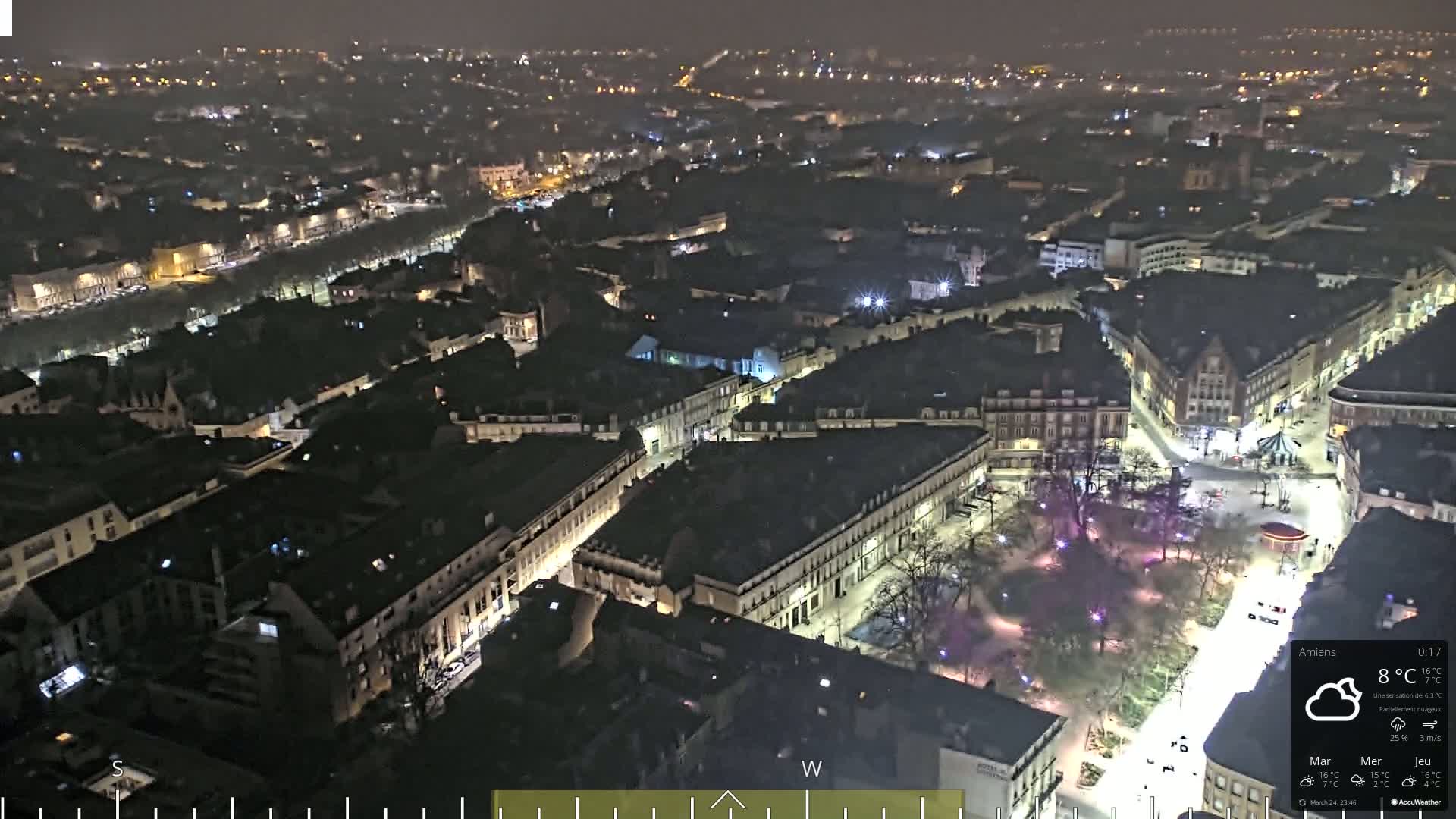 A nighttime aerial view of a city, showing dimly lit buildings and streets, with a partly cloudy sky.