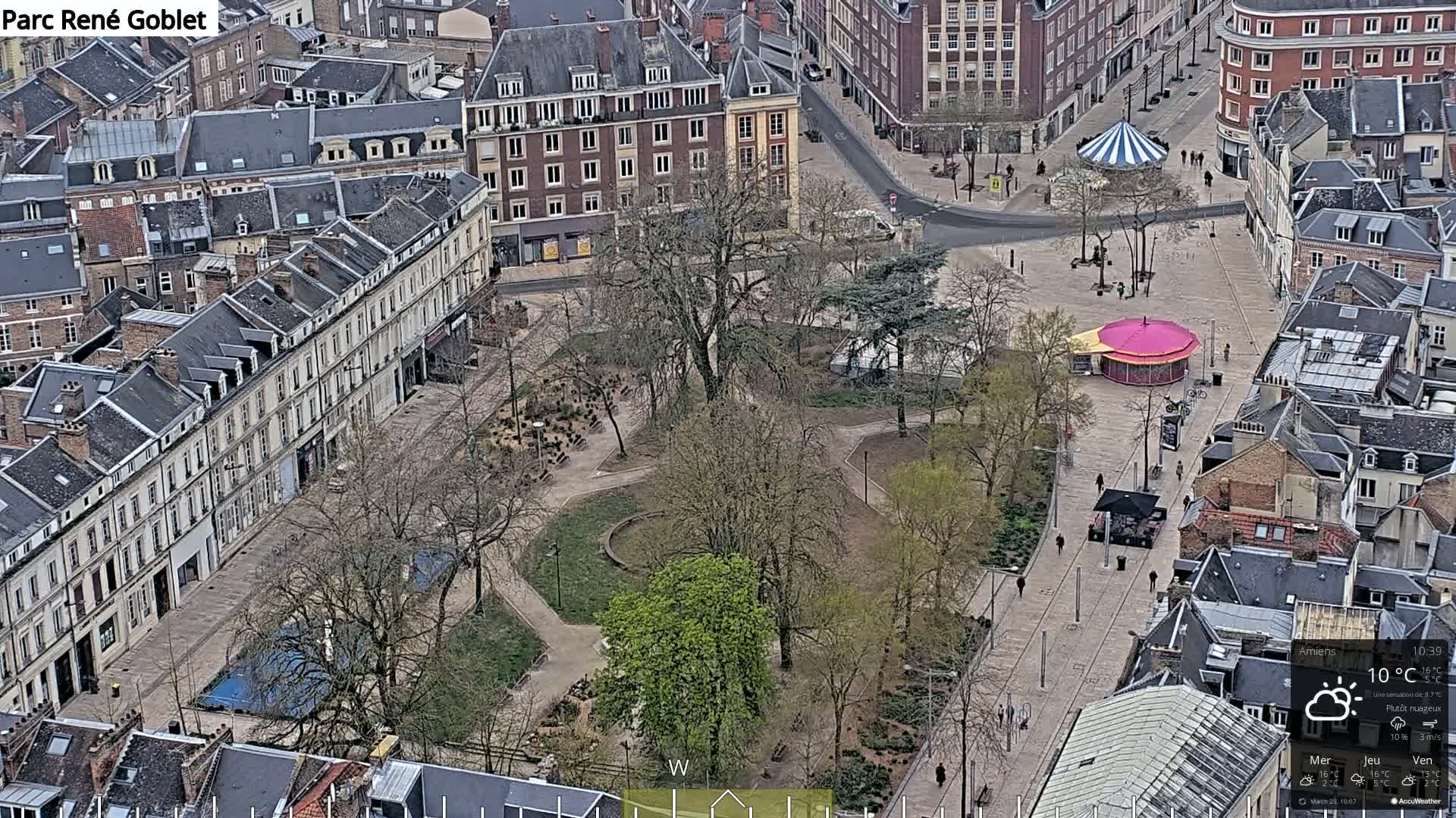 An aerial view shows a city square with a park in the center, surrounded by buildings under partly cloudy skies.