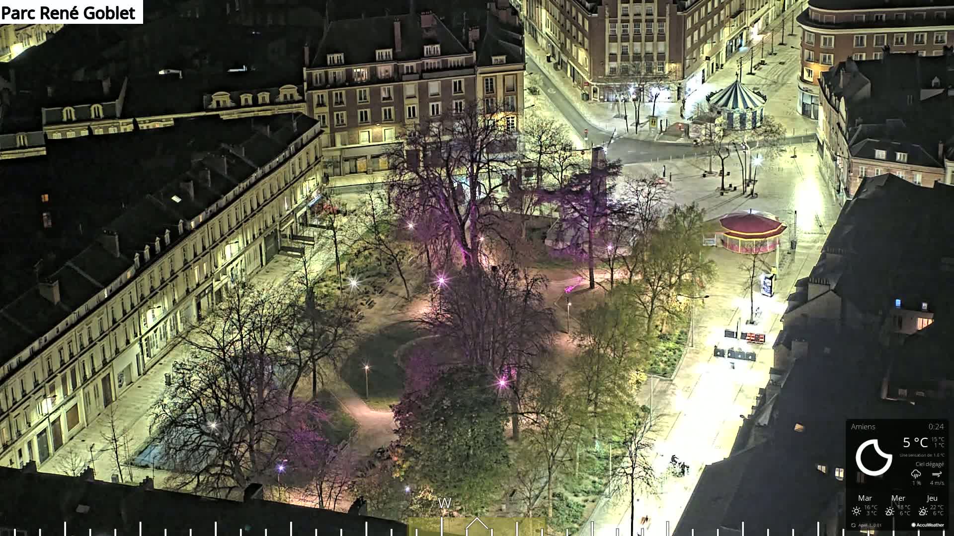 An aerial nighttime view of a park with trees and pathways, surrounded by city buildings under a clear sky.