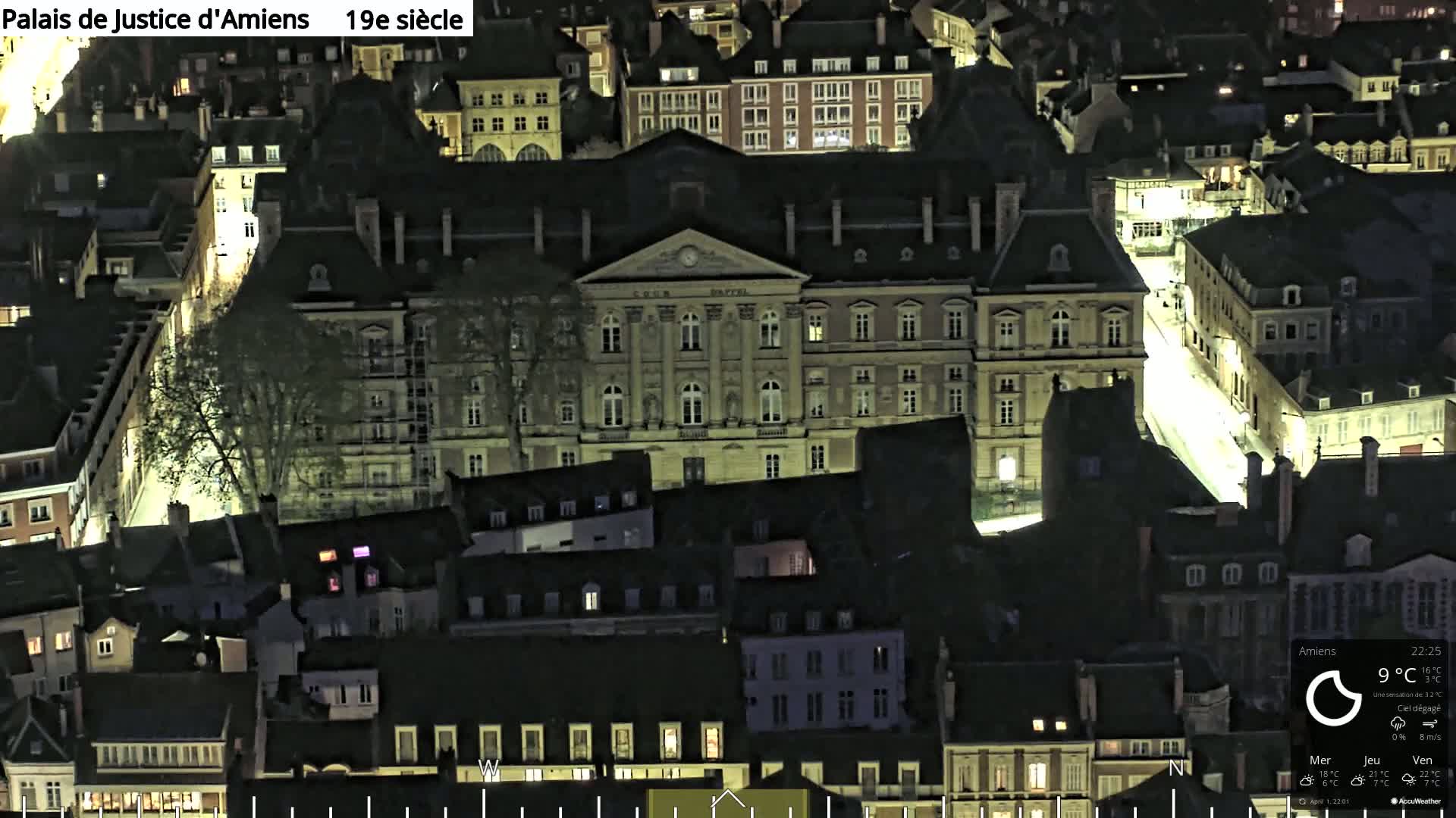 An aerial nighttime view of a city, showing a large, pale-colored building illuminated among many darker buildings, under a clear sky.