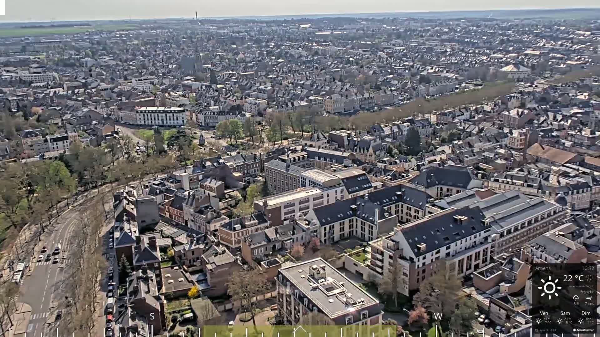 An aerial view of a densely populated city on a sunny day shows numerous buildings with gray slate roofs and tree-lined streets.