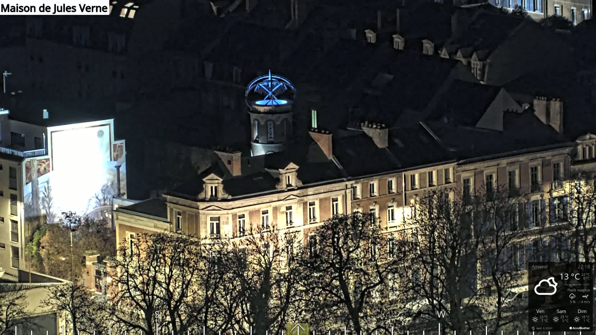 A nighttime aerial view of a city block featuring a building with a distinctive blue-lit cupola, surrounded by bare trees under a partly cloudy sky.