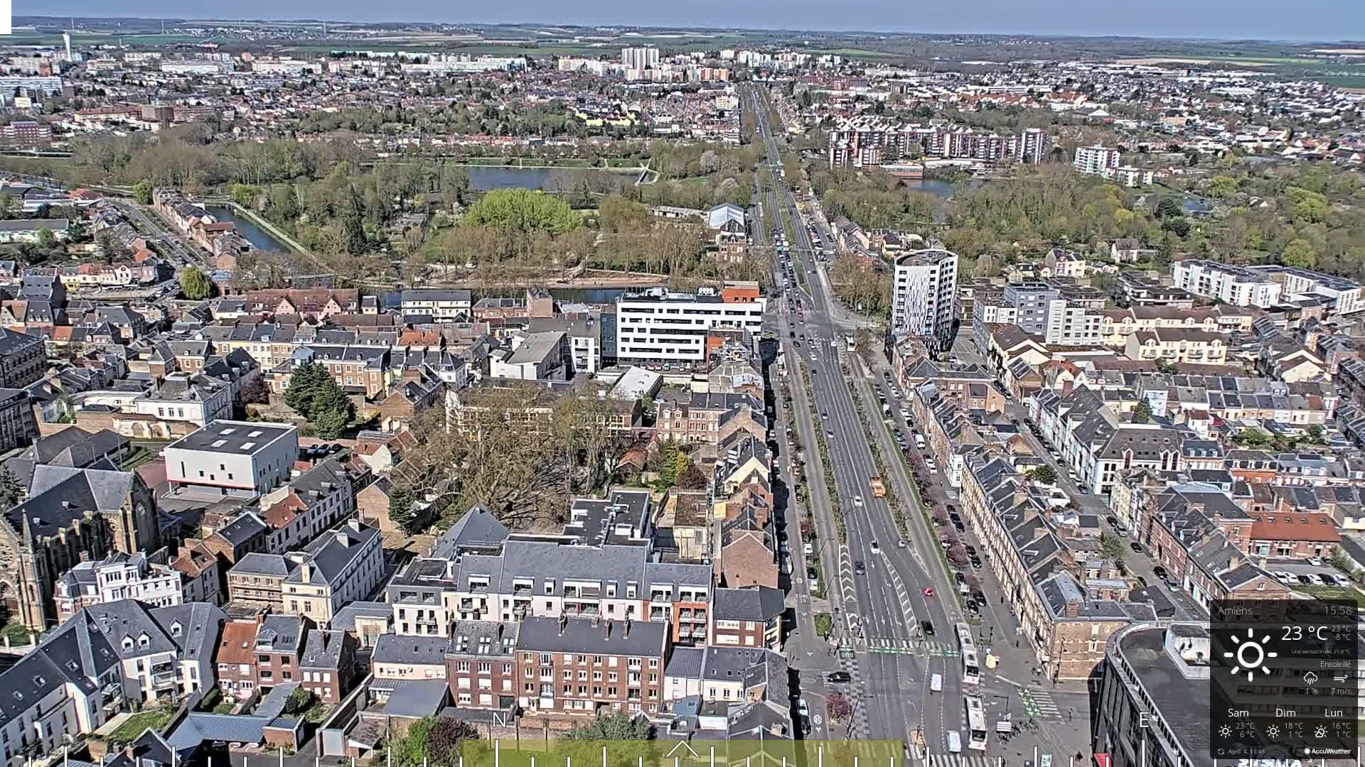 A high-angle, long shot of a city on a sunny day shows a main road cutting through a densely populated residential area, with greenery visible in the background.