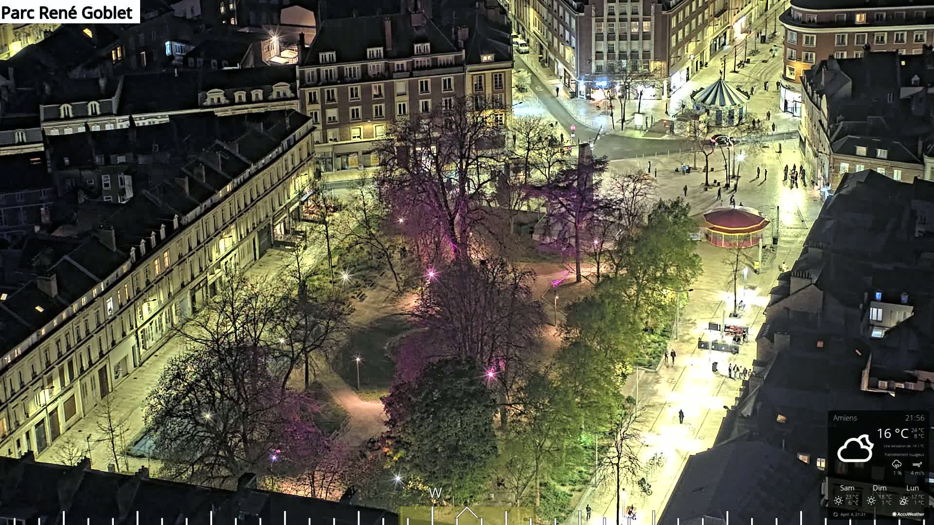 An aerial nighttime view of a city square with a park in the center, illuminated by streetlights, under partly cloudy skies.