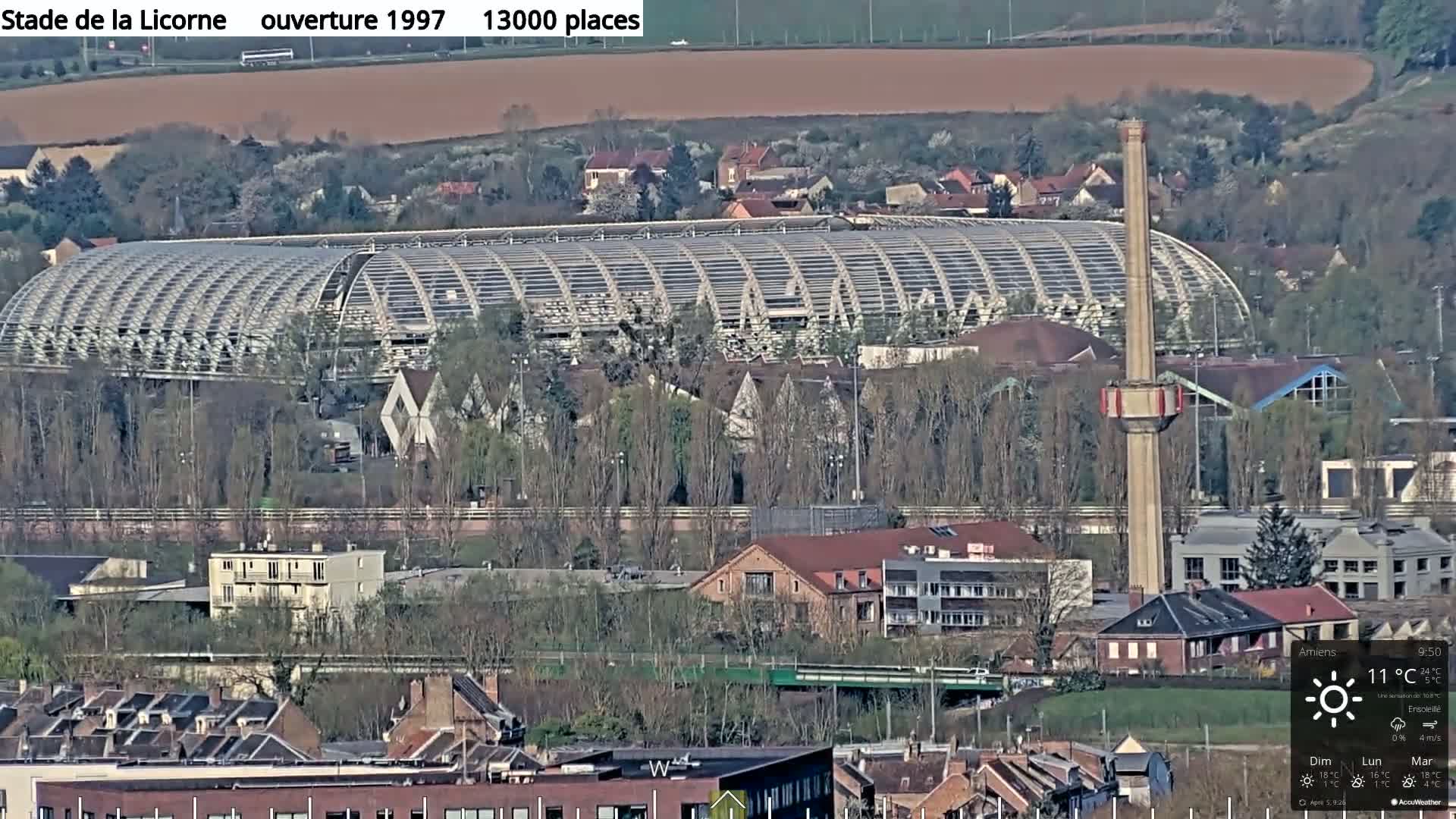 A large stadium with a metallic arched roof is situated amongst a town of low-rise buildings under a sunny sky.