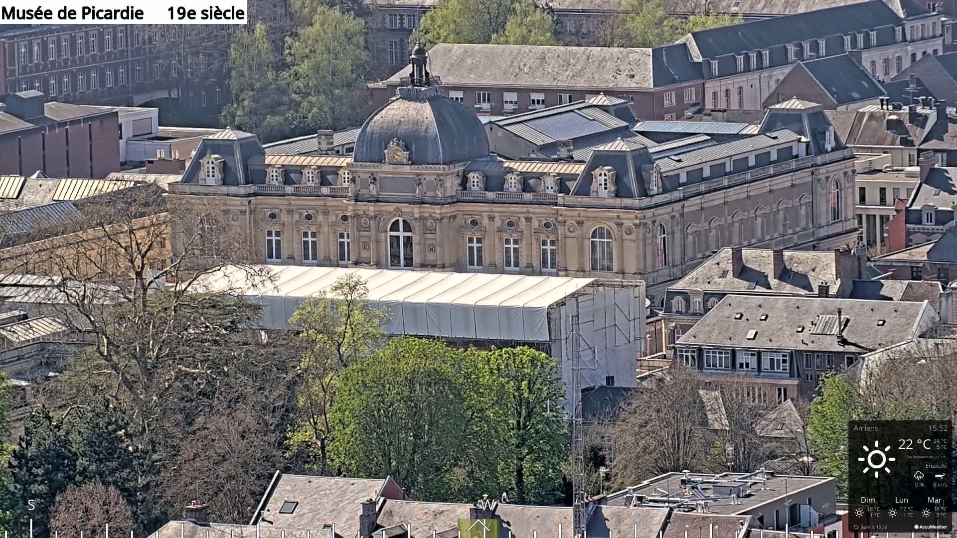 An aerial view of a large, light-beige building with a domed roof undergoing renovations under a sunny sky.