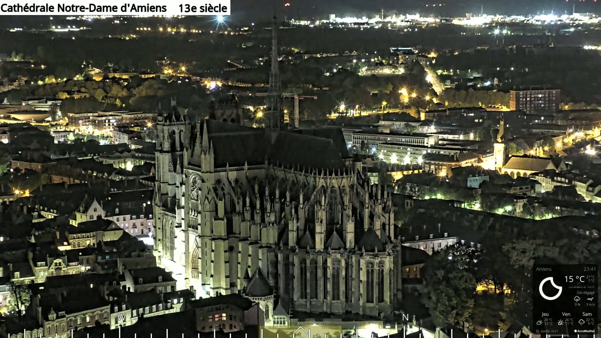 A large gothic cathedral is illuminated at night, surrounded by a city with many other buildings also lit up.