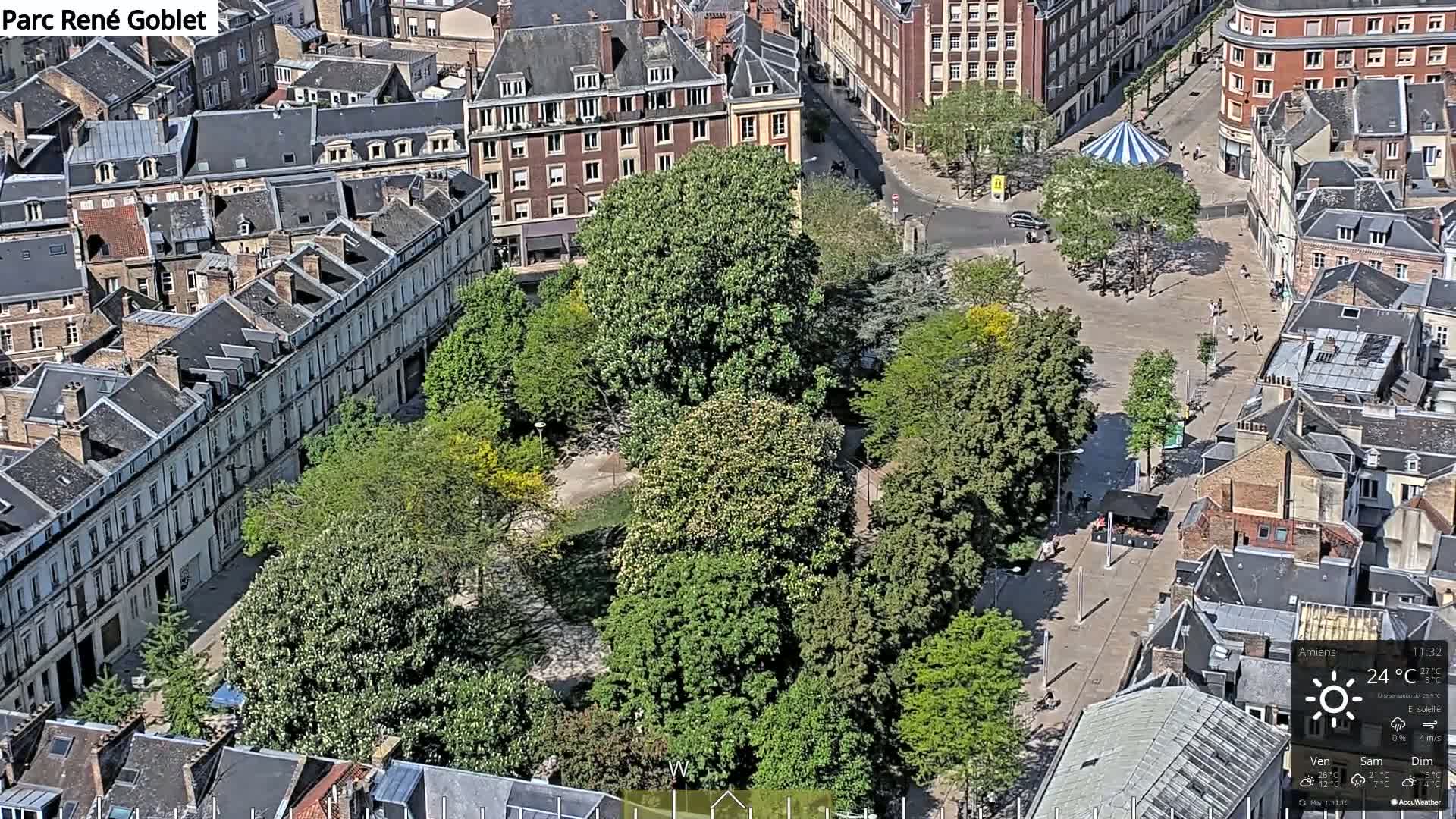 An aerial view shows a city square with a central park filled with lush green trees, surrounded by buildings on a sunny day.