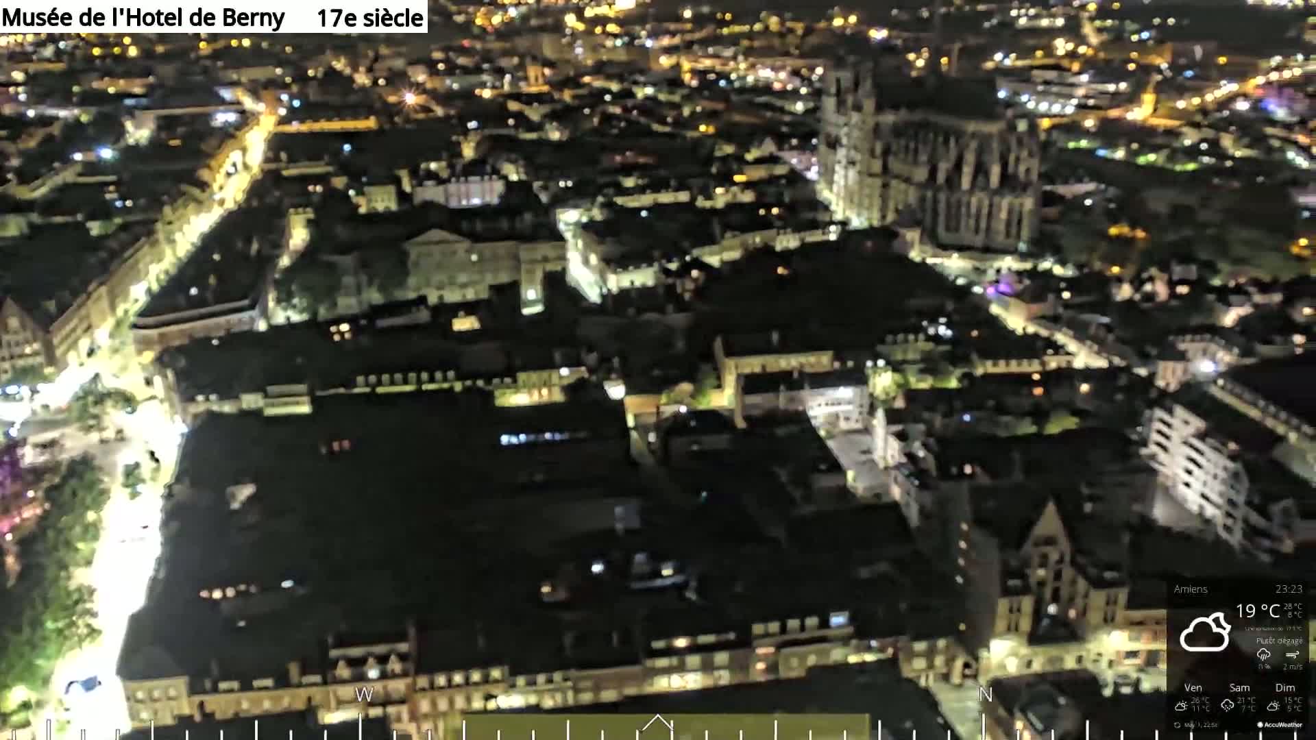 A nighttime aerial view of a city, showing illuminated streets and buildings, with a large cathedral visible in the center.