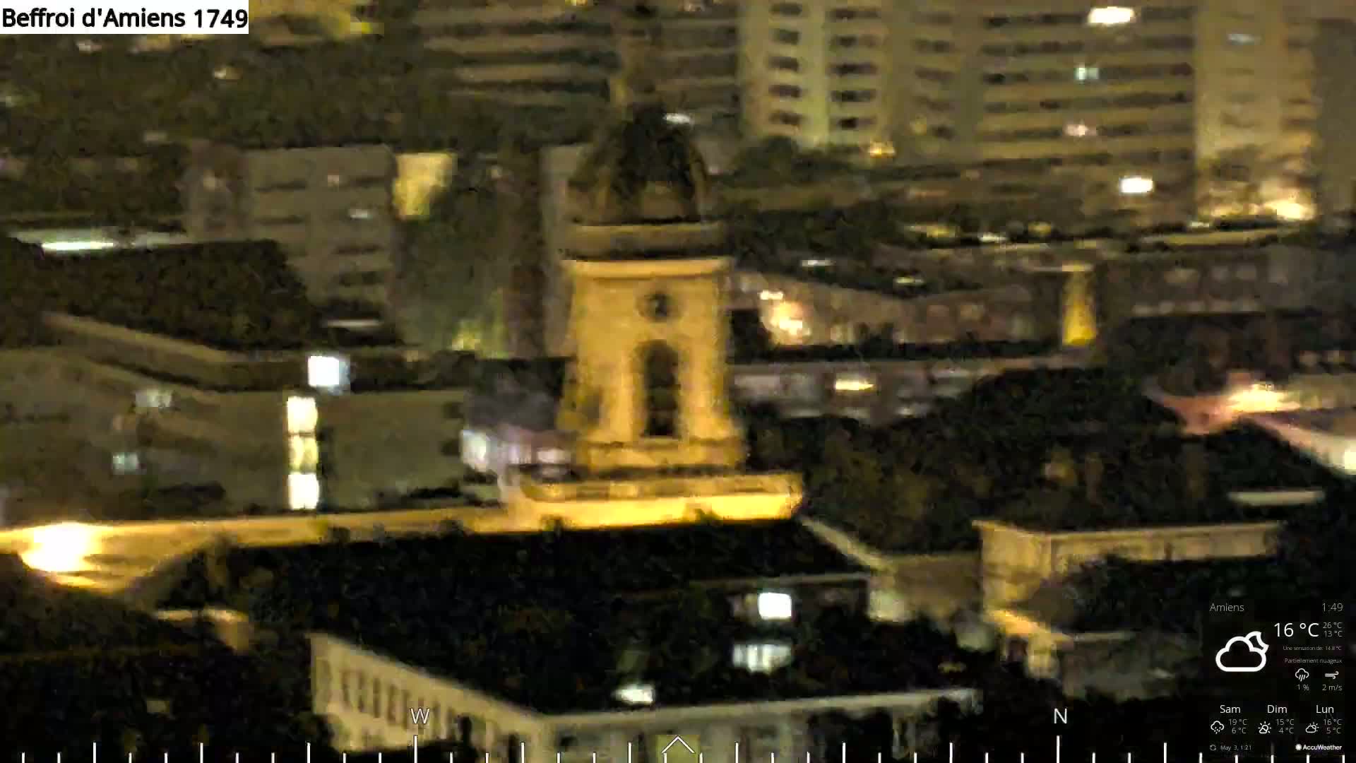 A nighttime, partly cloudy view of a city, showing a tall, illuminated stone structure among other buildings.
