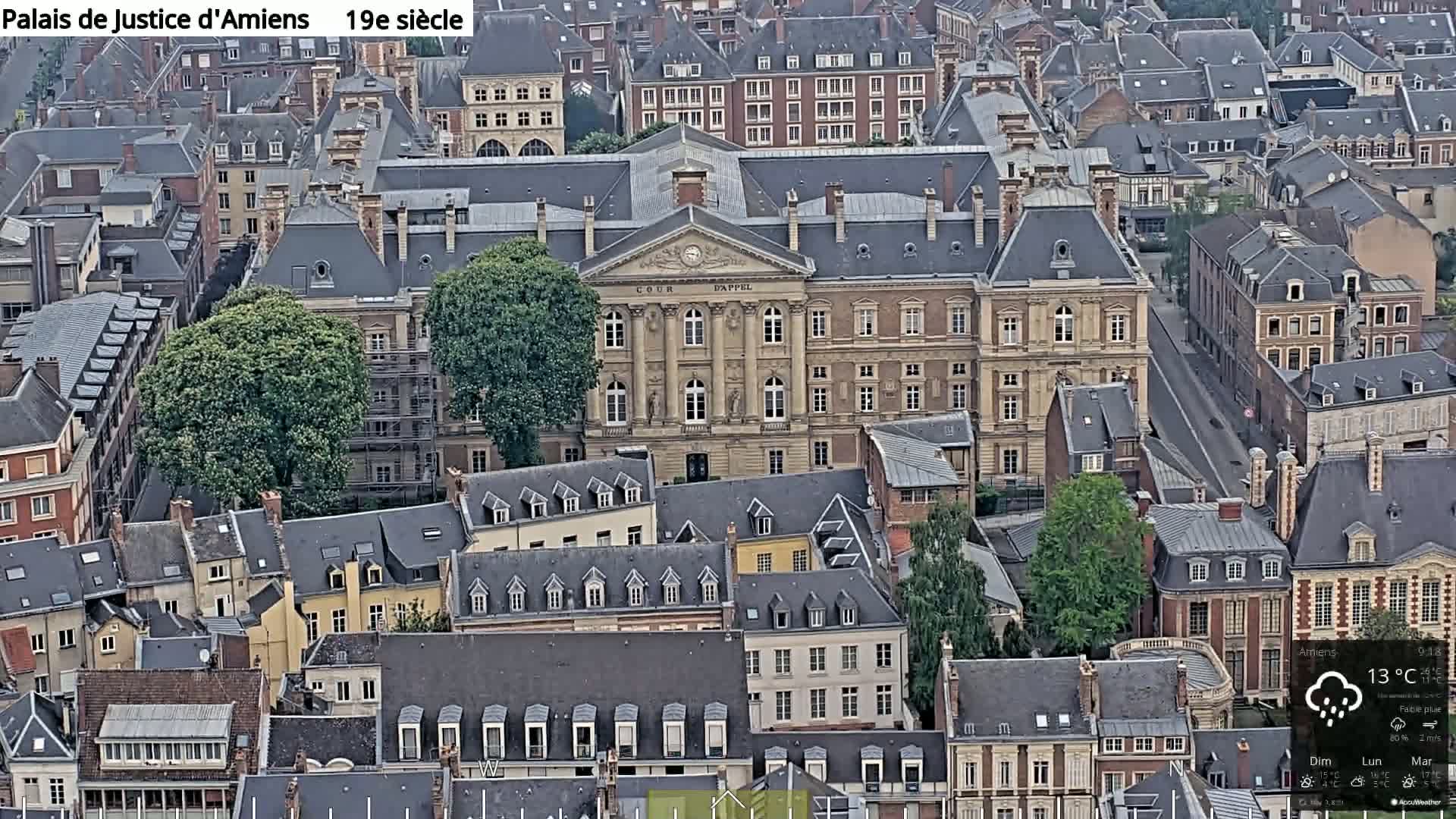 A high-angle view shows a city block of primarily gray-roofed buildings under a lightly overcast sky with a light rain.
