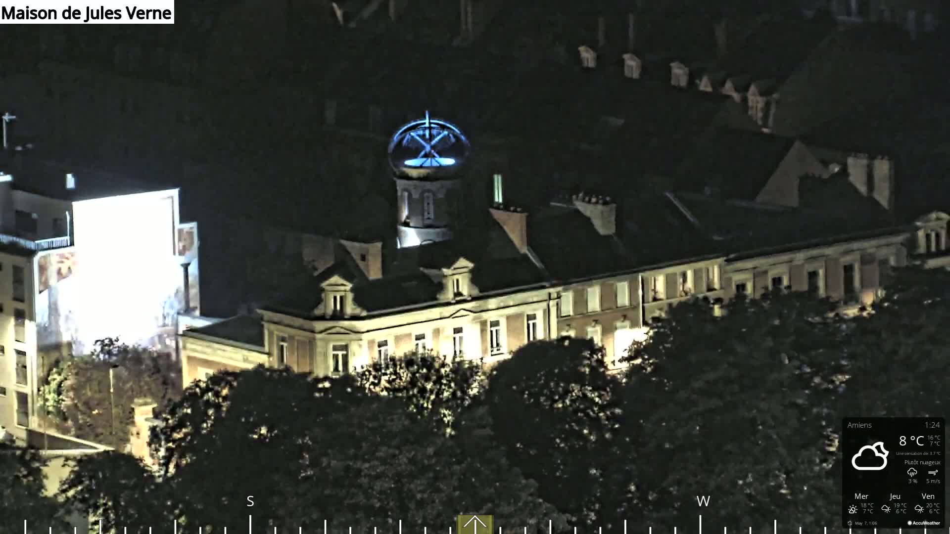 A nighttime aerial view of a large building complex with a tower illuminated in blue, surrounded by dark trees under partly cloudy skies.