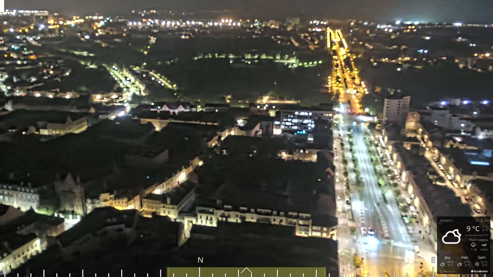 A nighttime, aerial view of a city with a long, brightly lit street running through it, under partly cloudy conditions.