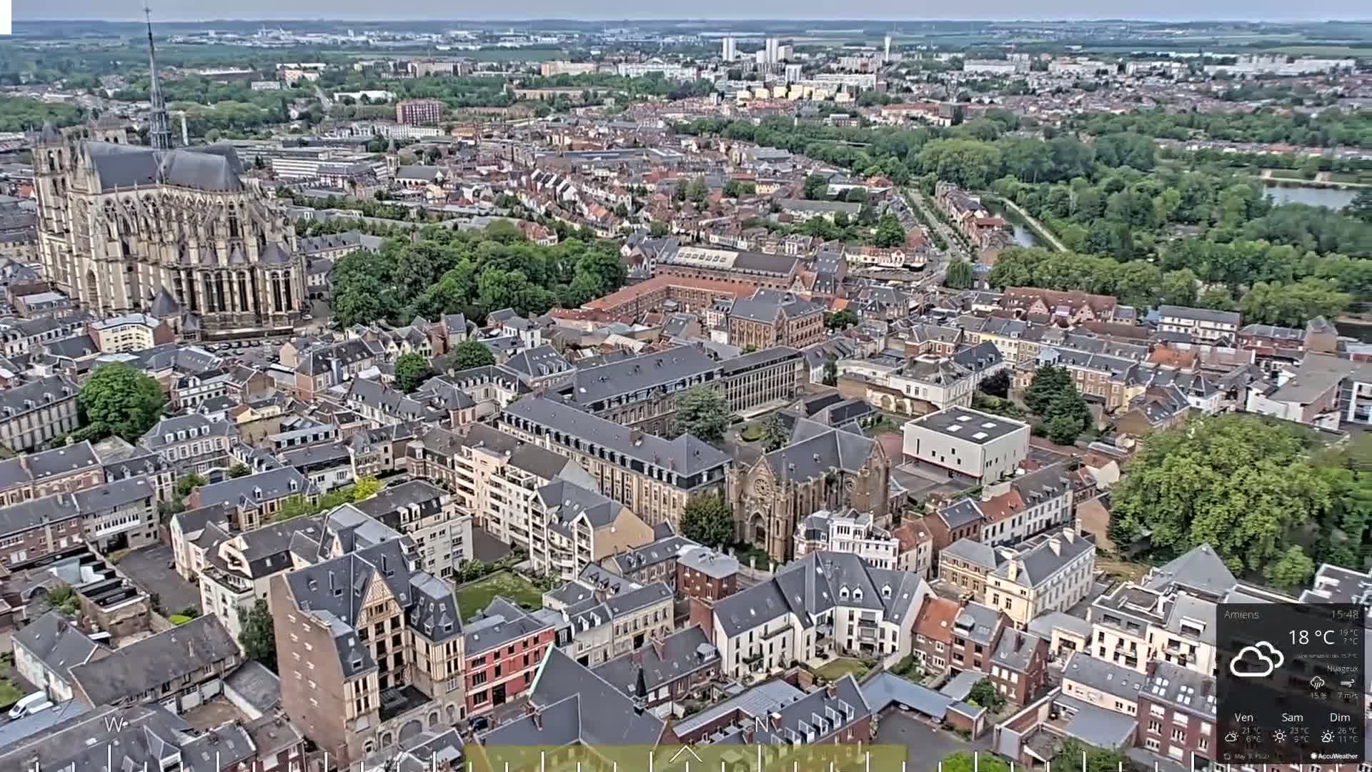 An aerial view shows a European city on a partly cloudy day, with a large cathedral prominent in the foreground.