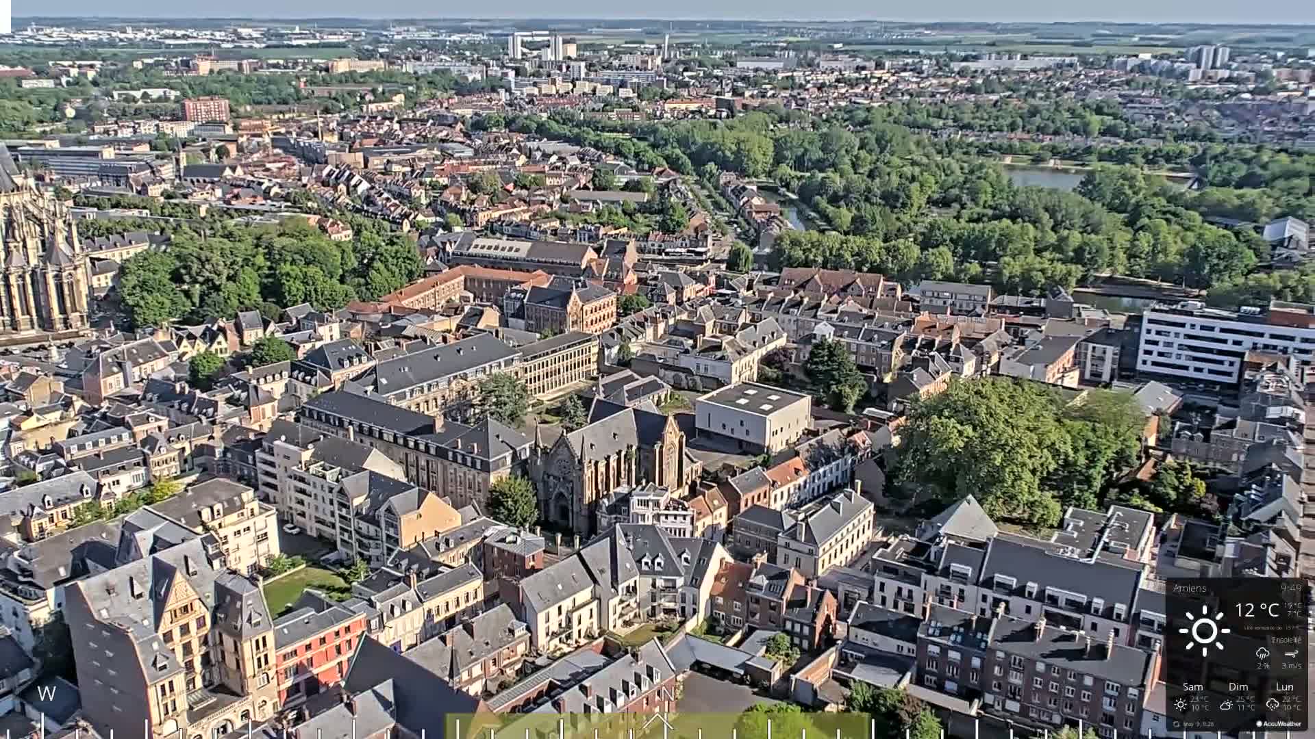 Here is a description of the image: An aerial view of a European city on a sunny day, showing a dense collection of buildings surrounded by green spaces and a river.