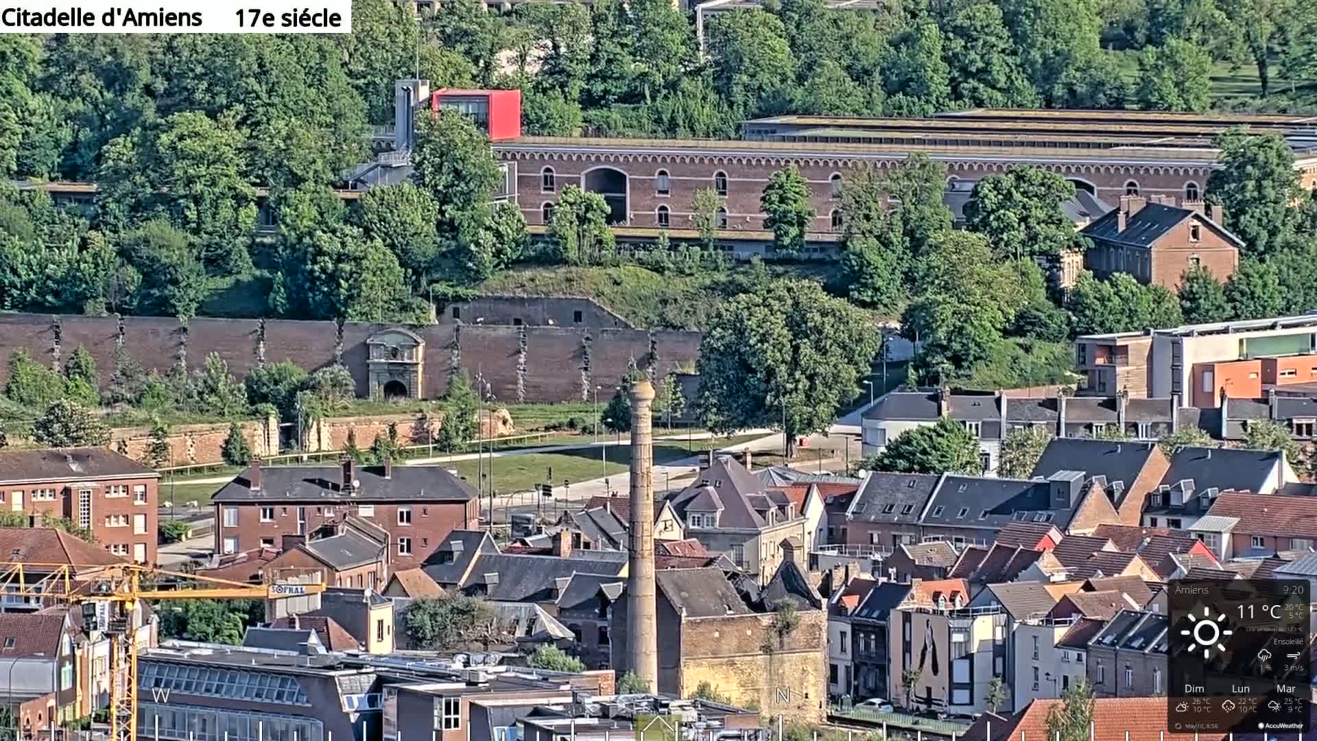 A sunny view of a European city with a brick citadel and tall chimney in the foreground, surrounded by numerous buildings and lush green trees.