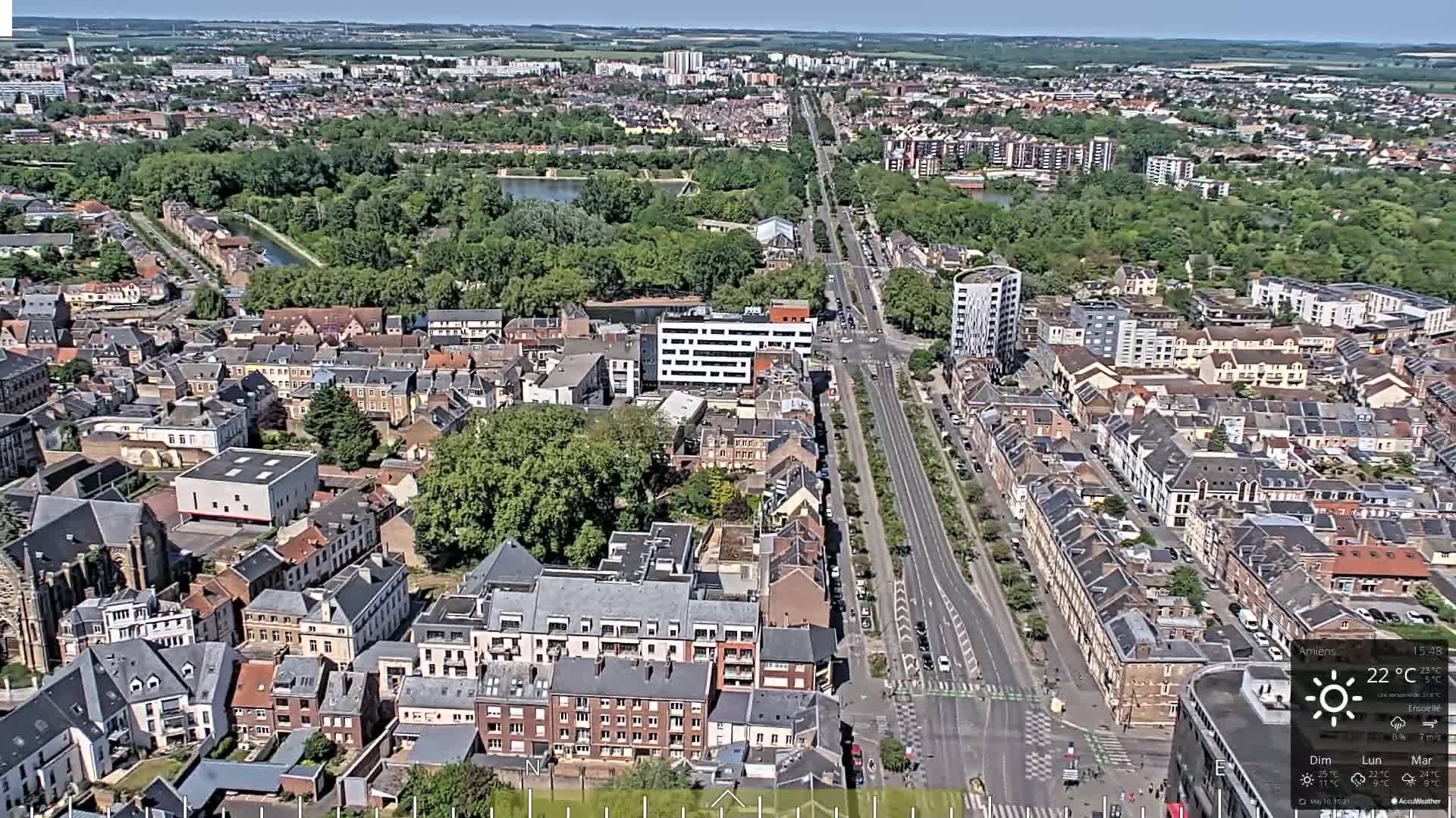 A high-angle, long shot of a city on a sunny day, showing a long road lined with buildings and trees, and extending to a wide expanse of greenery and further city in the distance.