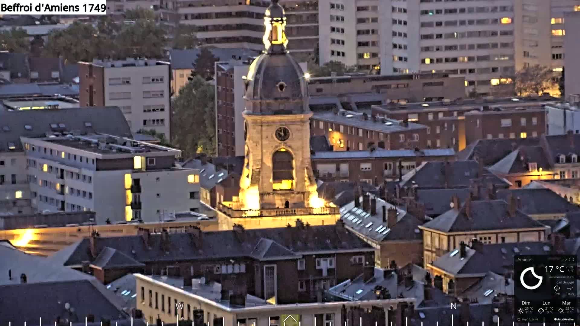 A brightly lit, ornate clock tower stands in the center of a city at dusk, surrounded by dark-roofed buildings under a clear sky.