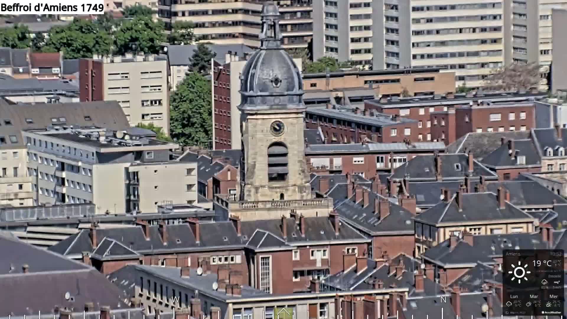 A clock tower stands amidst a densely packed cityscape of gray and red-roofed buildings under a sunny sky.