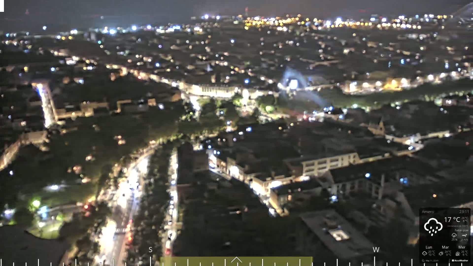 A nighttime aerial view of a city with lightly trafficked roads and dimly lit buildings under a slightly rainy sky.