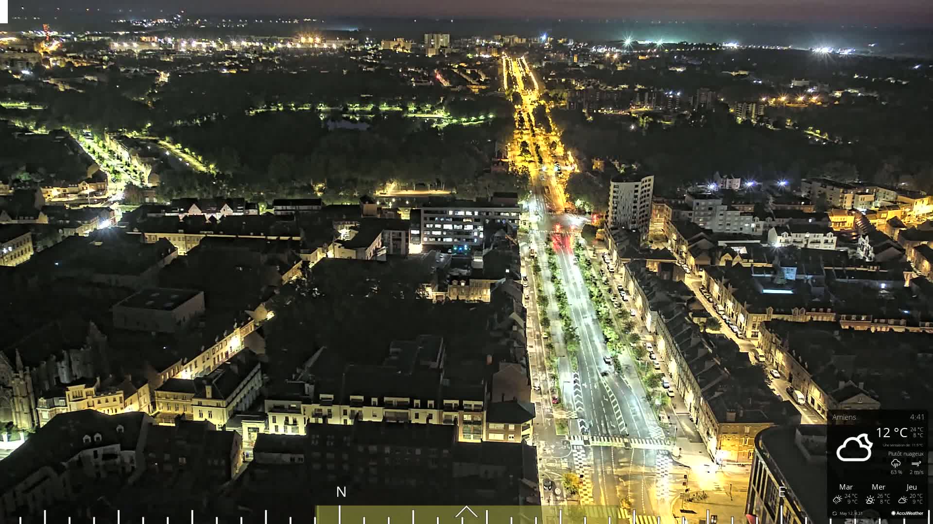 A brightly lit city street at night, surrounded by residential buildings and dark green spaces, under a partly cloudy sky.