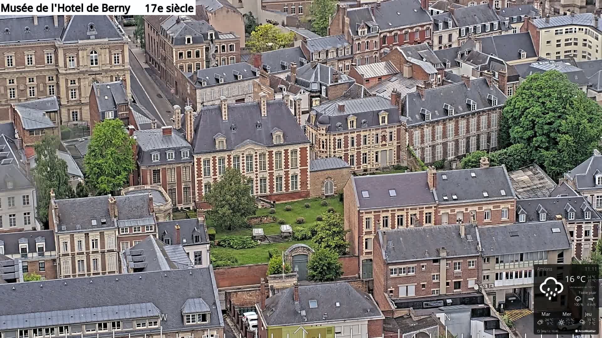 An aerial view shows a large brick building surrounded by smaller buildings in a city under lightly cloudy skies with a chance of rain.