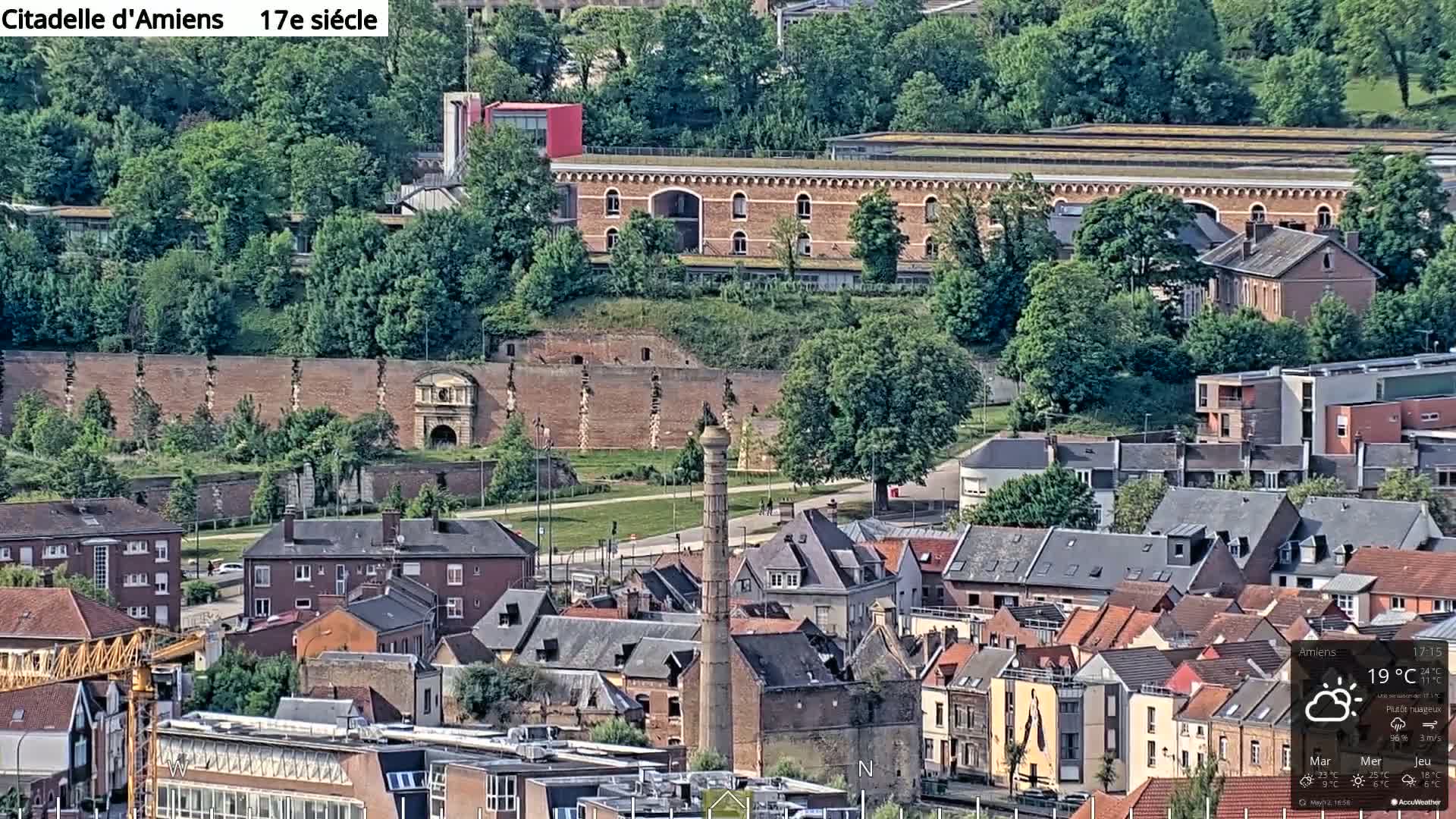 A high-angle, long shot of a town with a brick citadel and many buildings under a partly sunny sky.