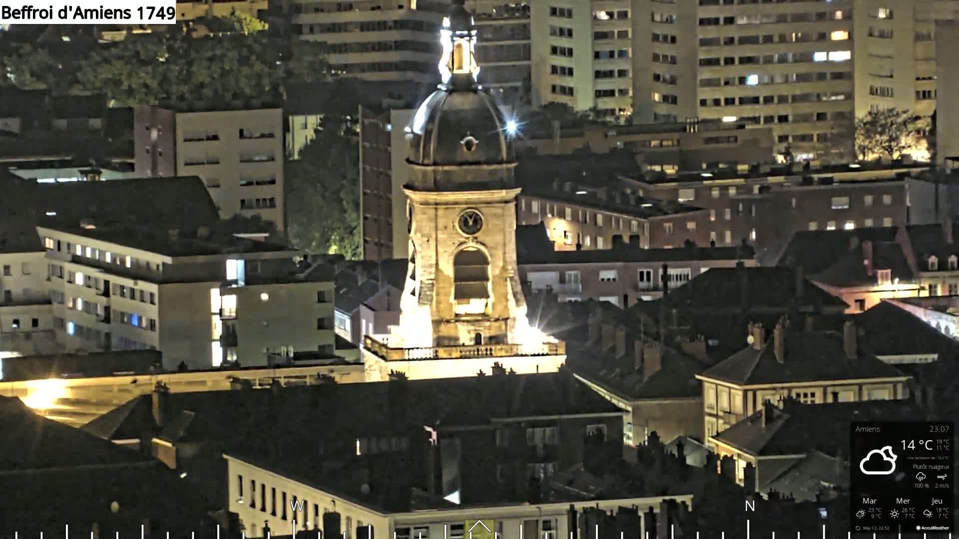 A brightly lit clock tower stands prominently among a cityscape at night, under partly cloudy skies.