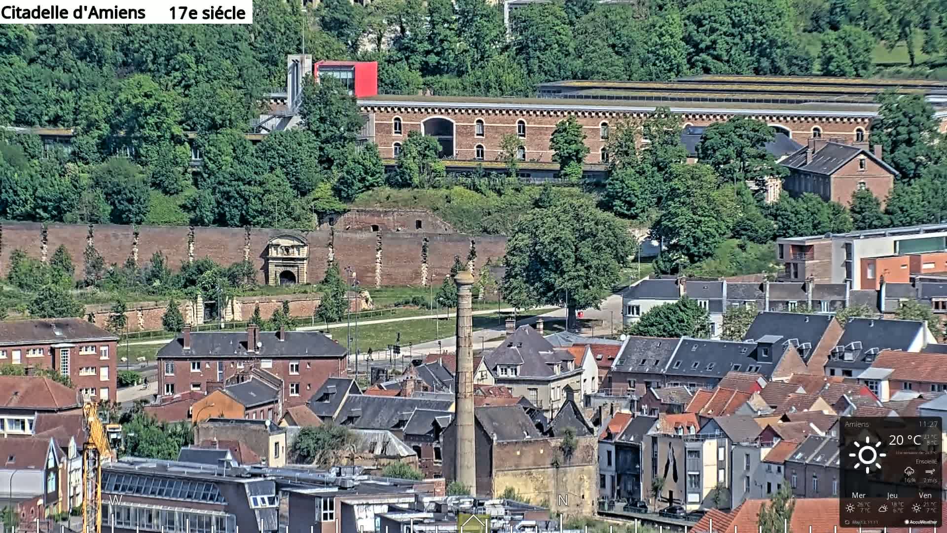 An elevated view of a European town on a sunny day, featuring a large brick wall and gateway, surrounded by buildings of varying heights and architectural styles.