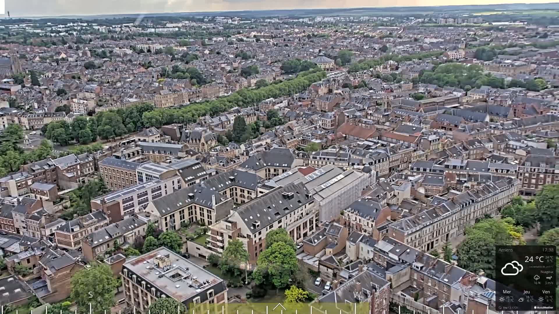 An aerial view of a densely packed city on a cloudy day shows numerous buildings and streets with a band of green trees.