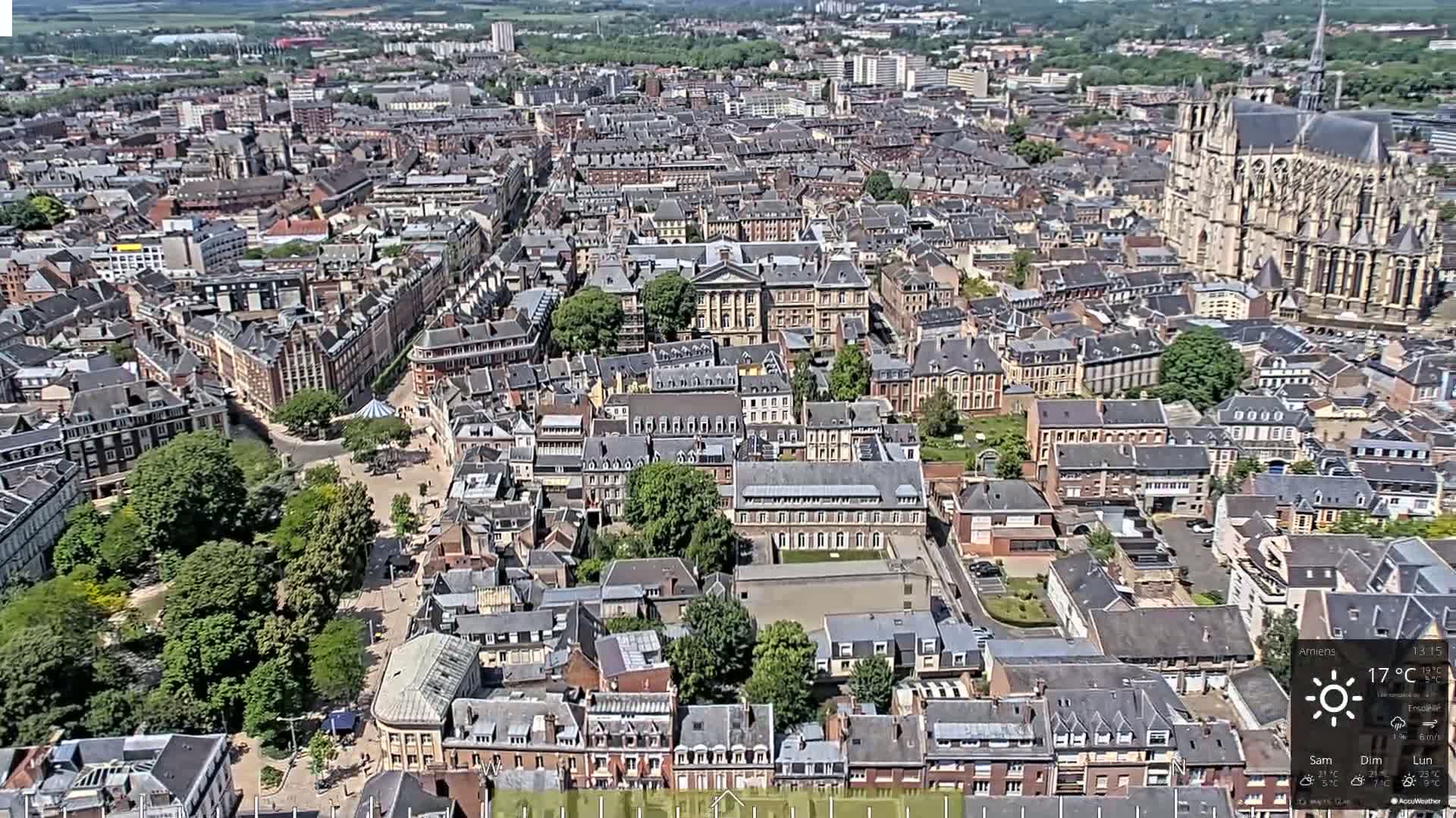 An aerial view of a European city on a sunny day shows a dense collection of buildings, a large cathedral, and a central square with green trees.