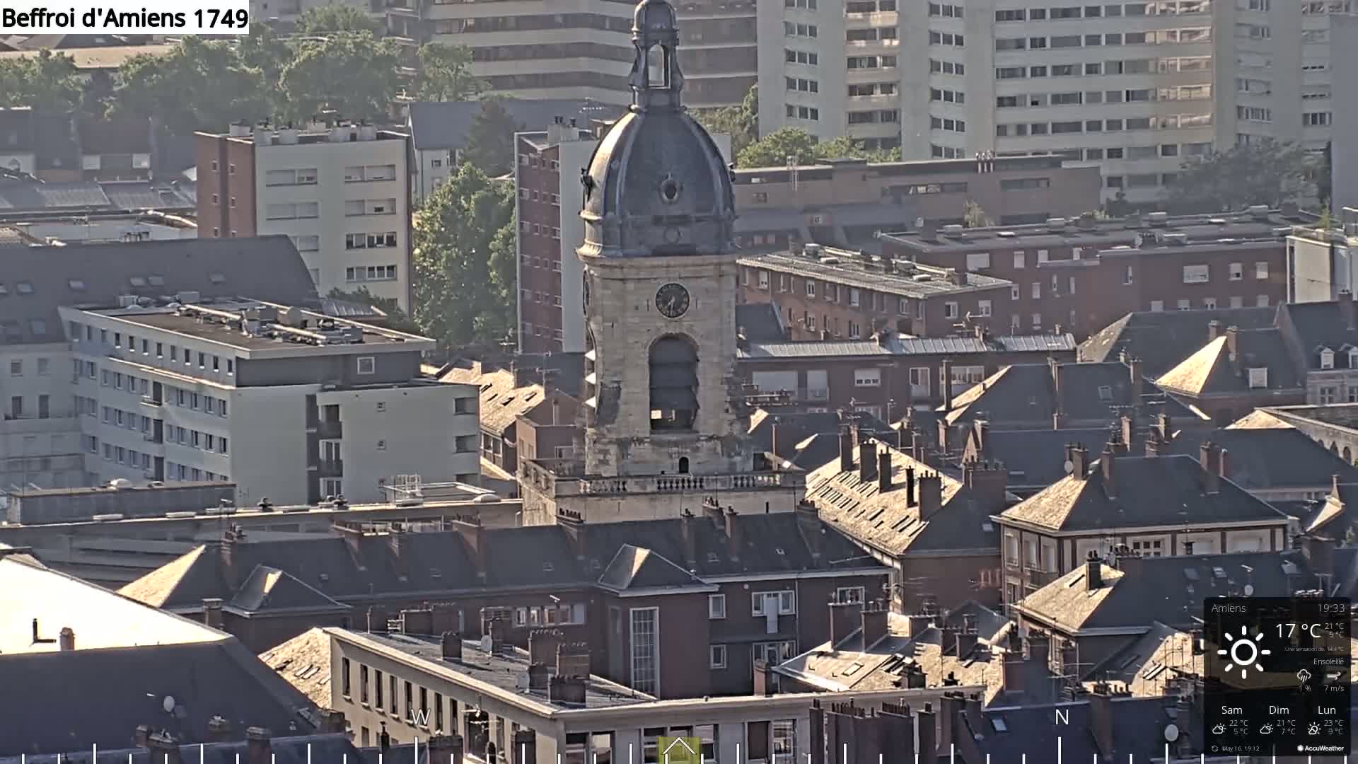 A high-angle, long shot of a city on a sunny day, centered on a tall stone tower with a domed roof, surrounded by many buildings with dark-colored pitched roofs.