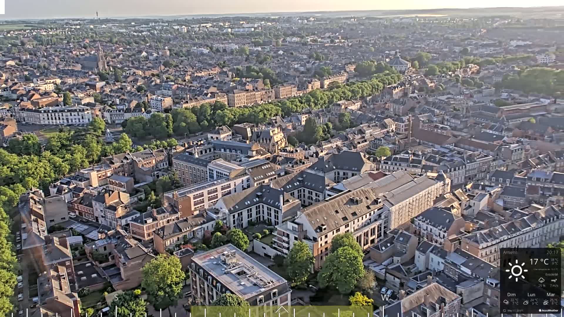 An aerial view of a densely populated city on a sunny day shows many closely packed buildings and green spaces.